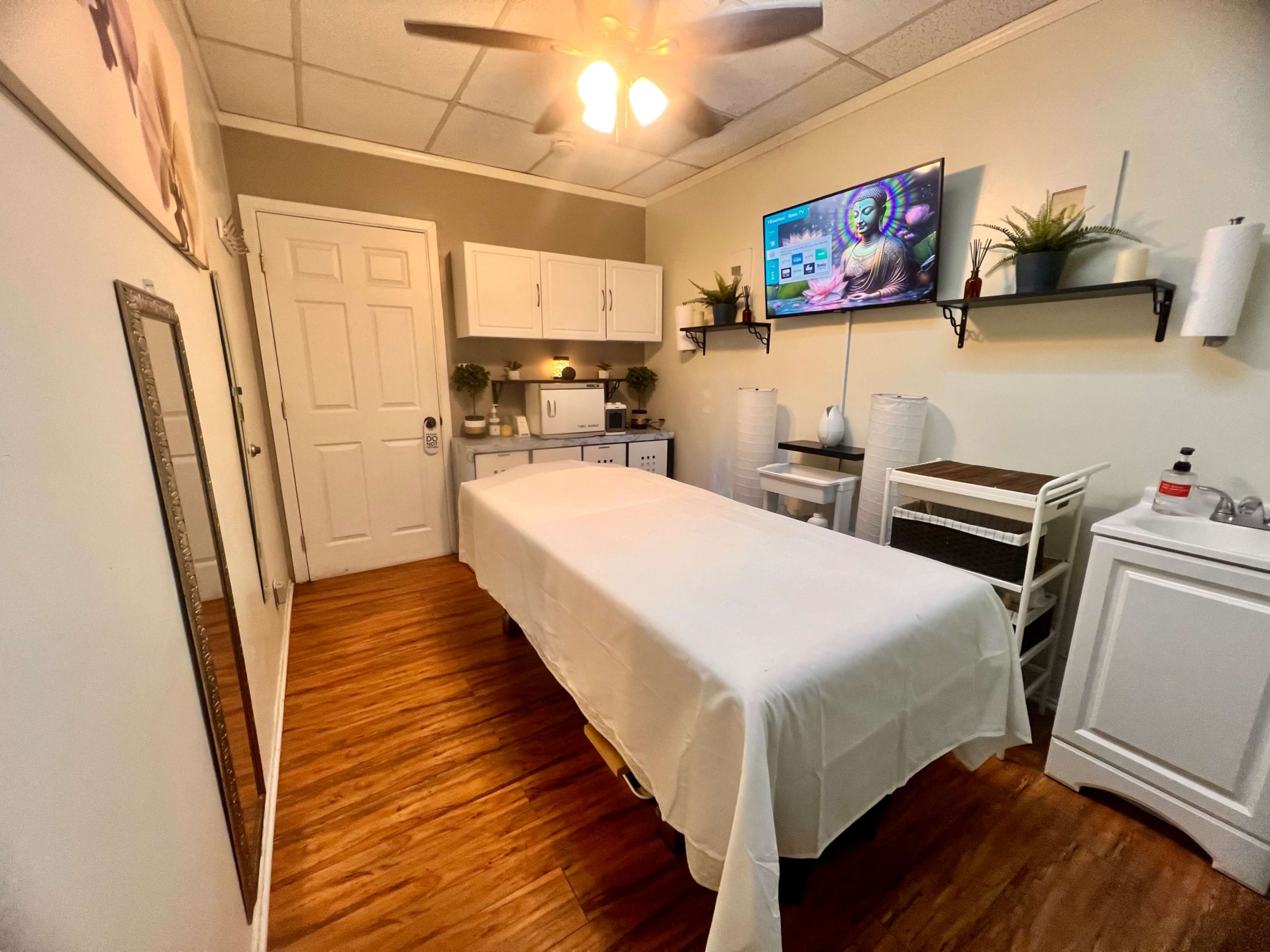 The image shows a treatment room with a massage table covered in a white sheet, shelves on the walls, and a television mounted above a cabinet.