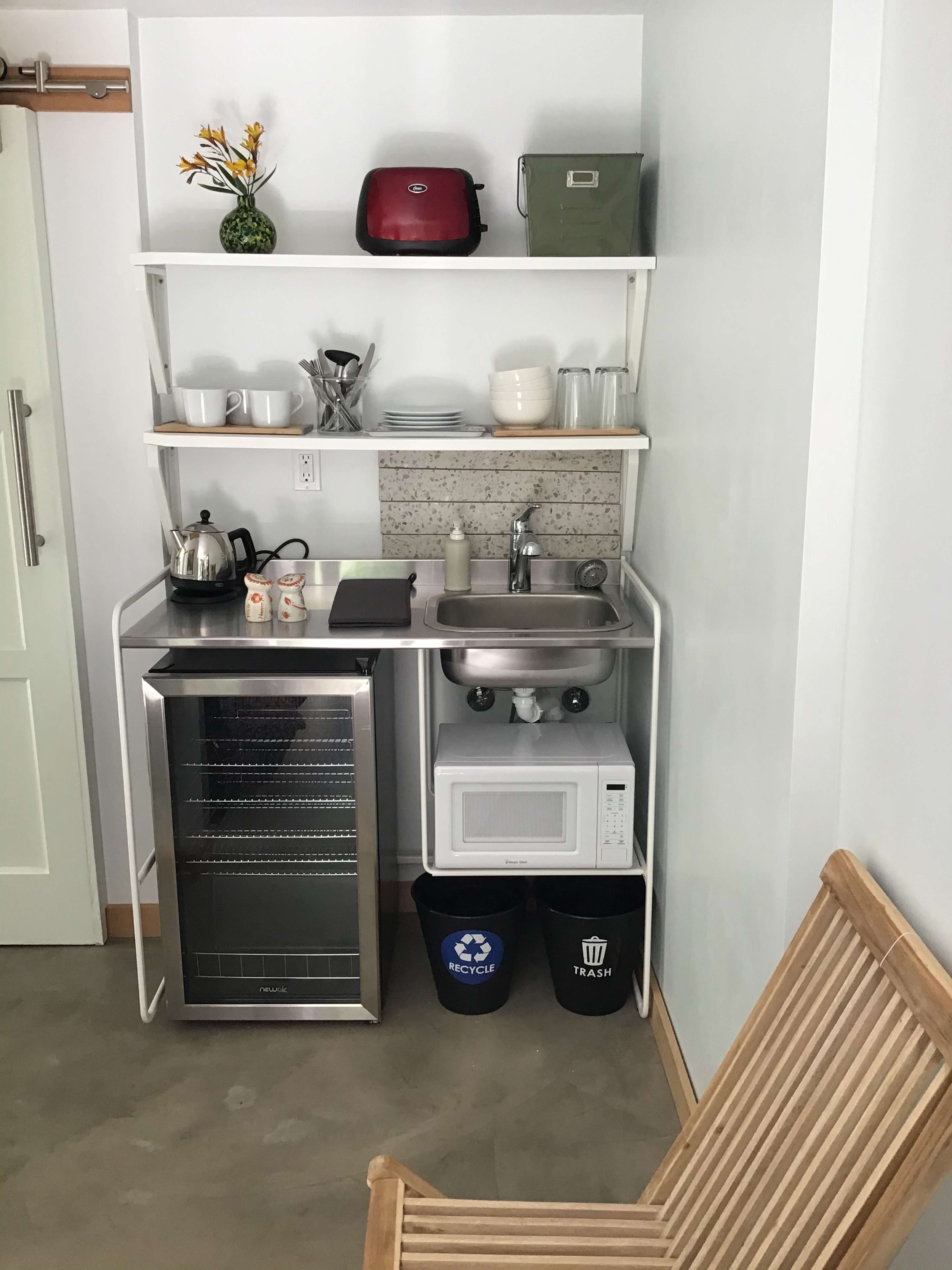 The image shows a compact kitchen area with a small refrigerator, sink, microwave, and various kitchen items arranged on open shelves.