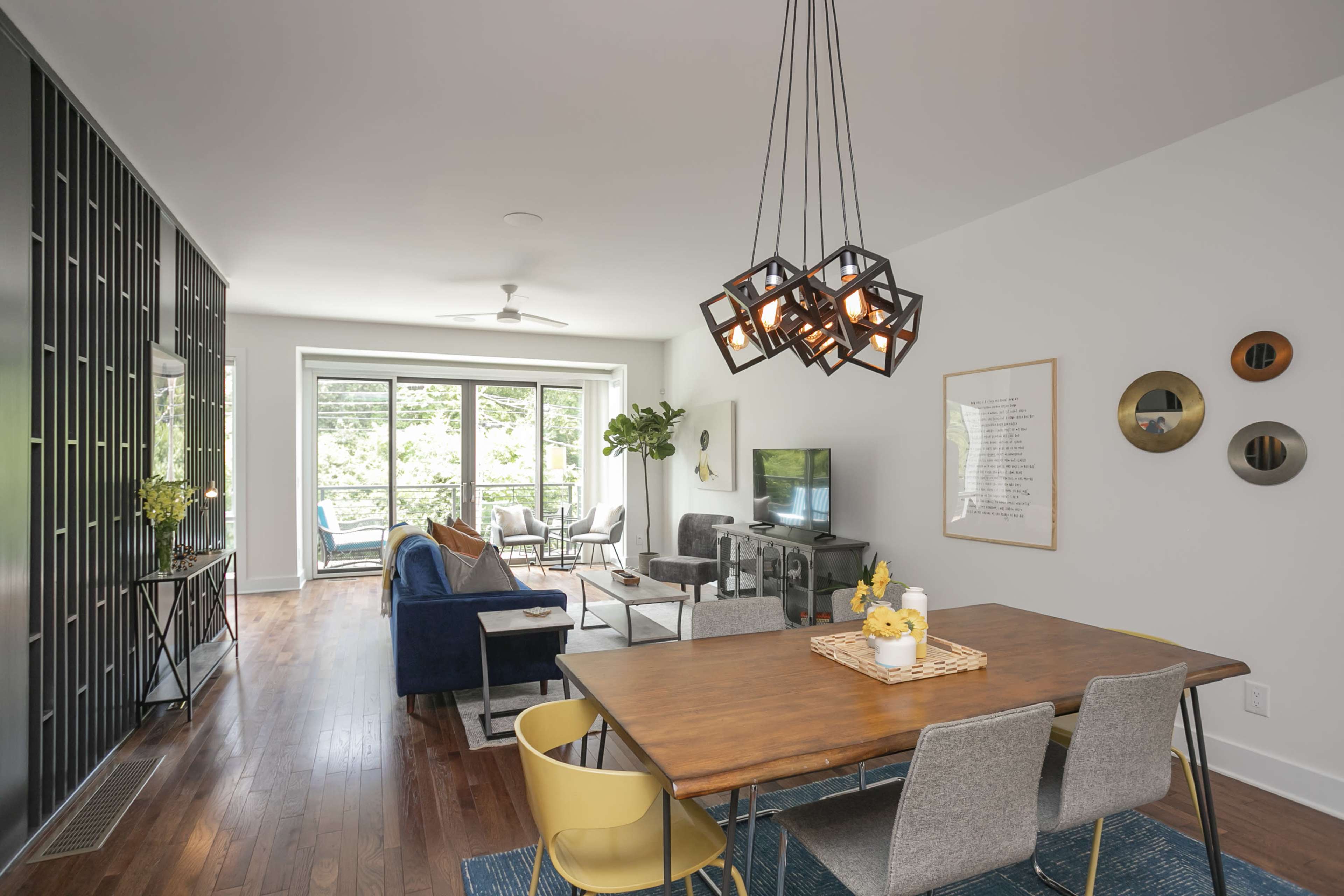 A modern living space features a dining area with a wooden table and chairs, a lounge area with a sofa and television, and large windows overlooking greenery.