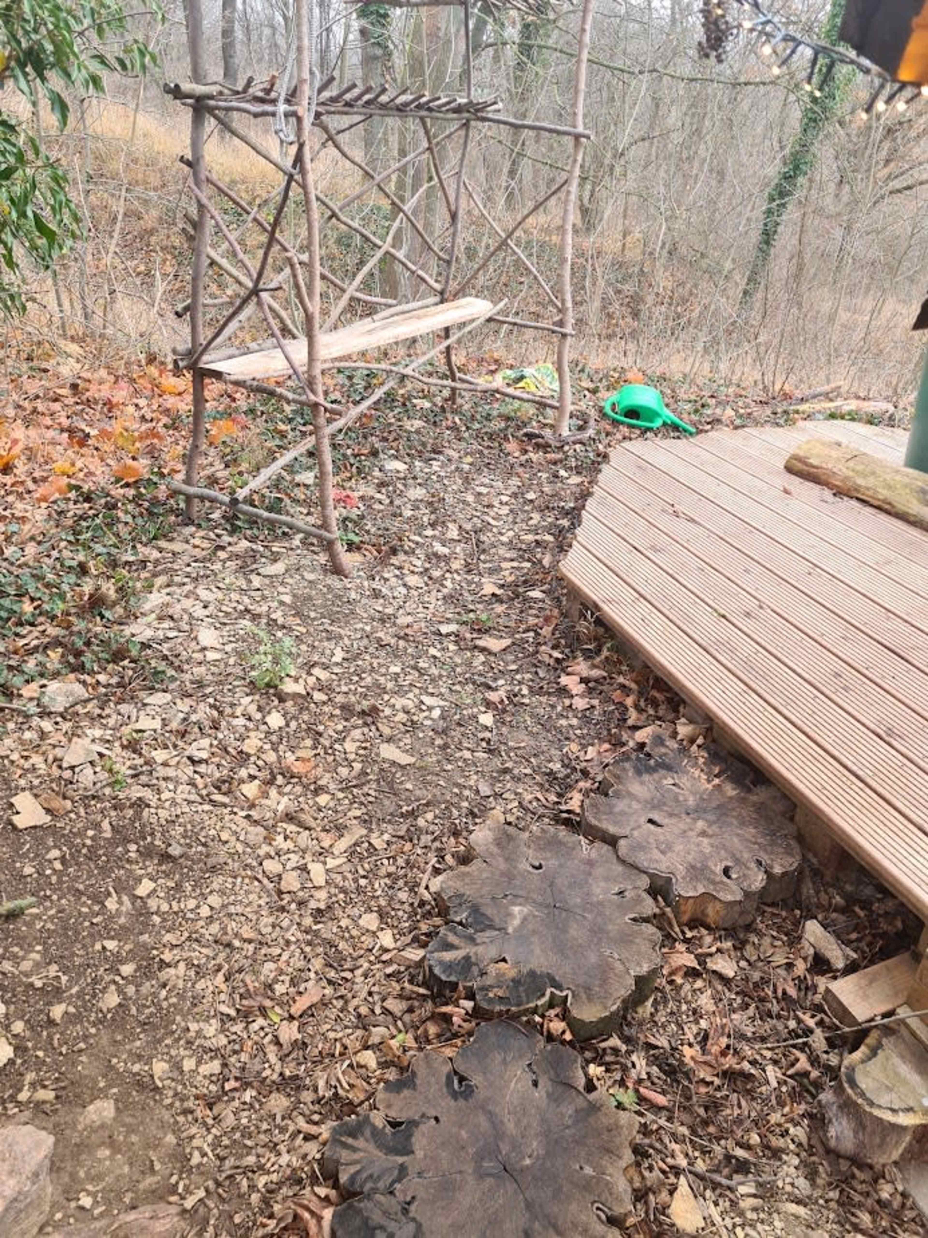 The image shows a rustic wooden bench made from branches positioned near a gravel path leading to a deck, with log slices forming stepping stones.