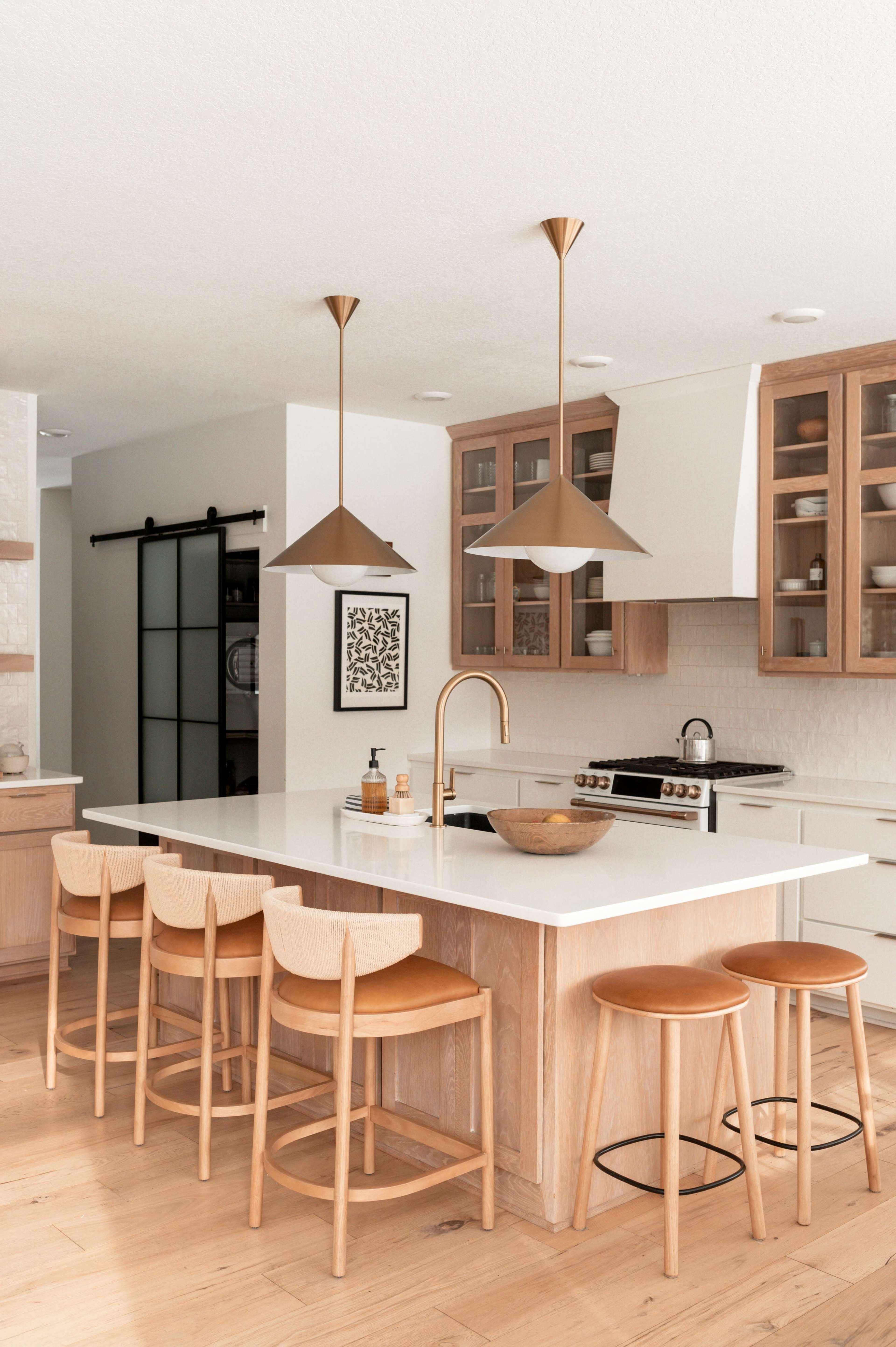 A bright kitchen features an island with wooden stools, pendant lights, and modern appliances, alongside glass-fronted cabinets.