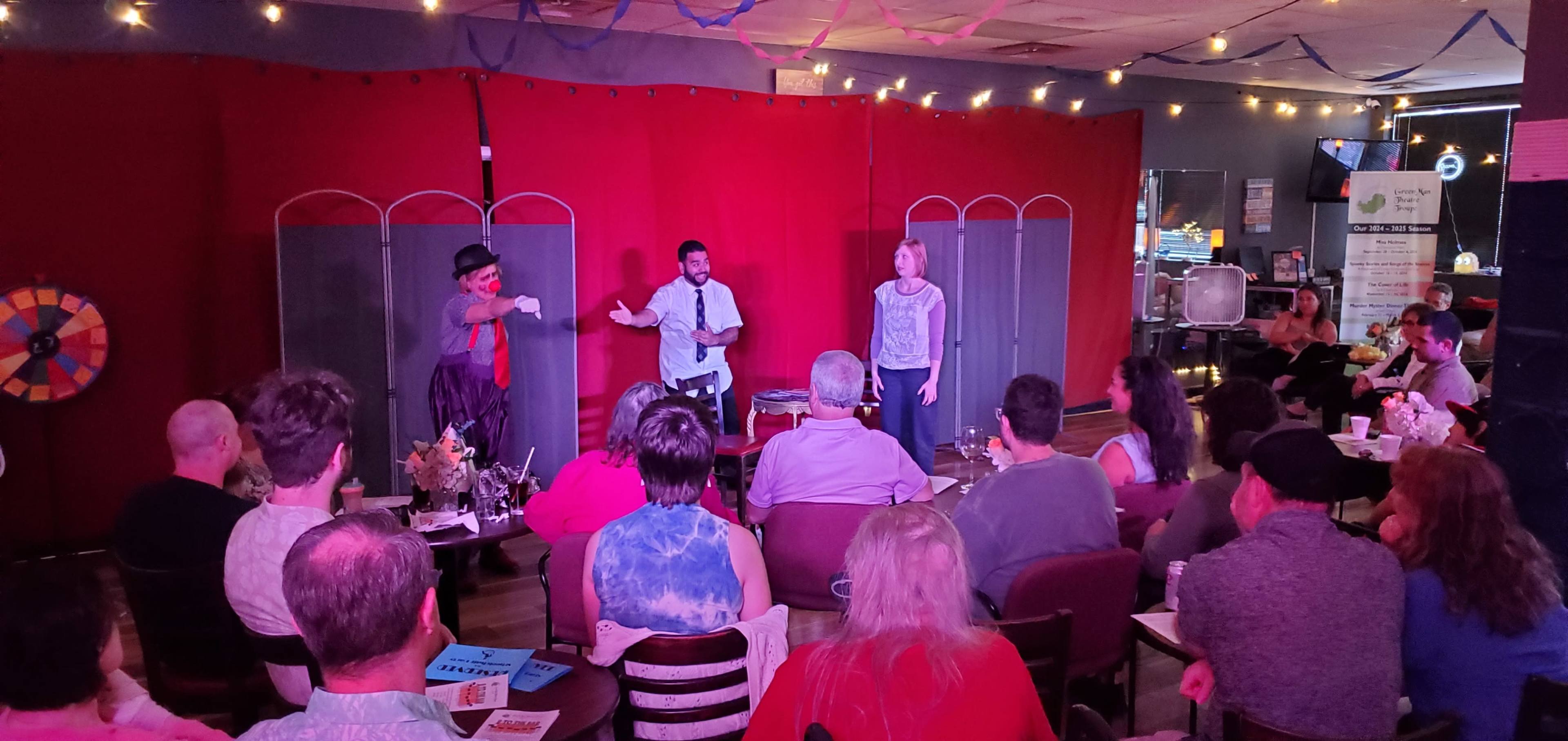 A small audience watches a performance on stage featuring three entertainers in a cozy venue adorned with colorful decorations.