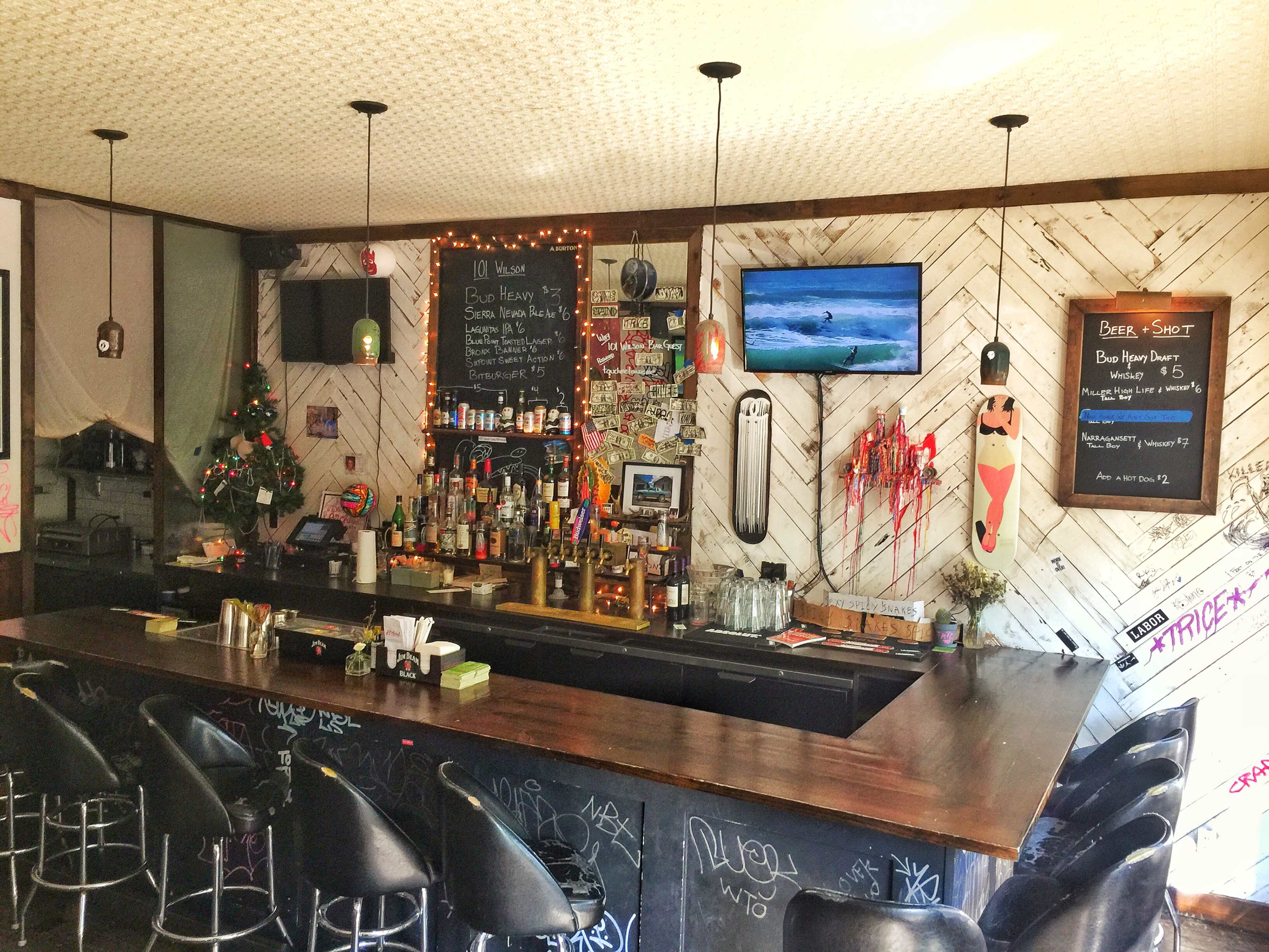 A bar area with a dark wood counter, shelves of bottles, and a TV displaying a surfing scene, surrounded by festive decorations and graffiti on the walls.