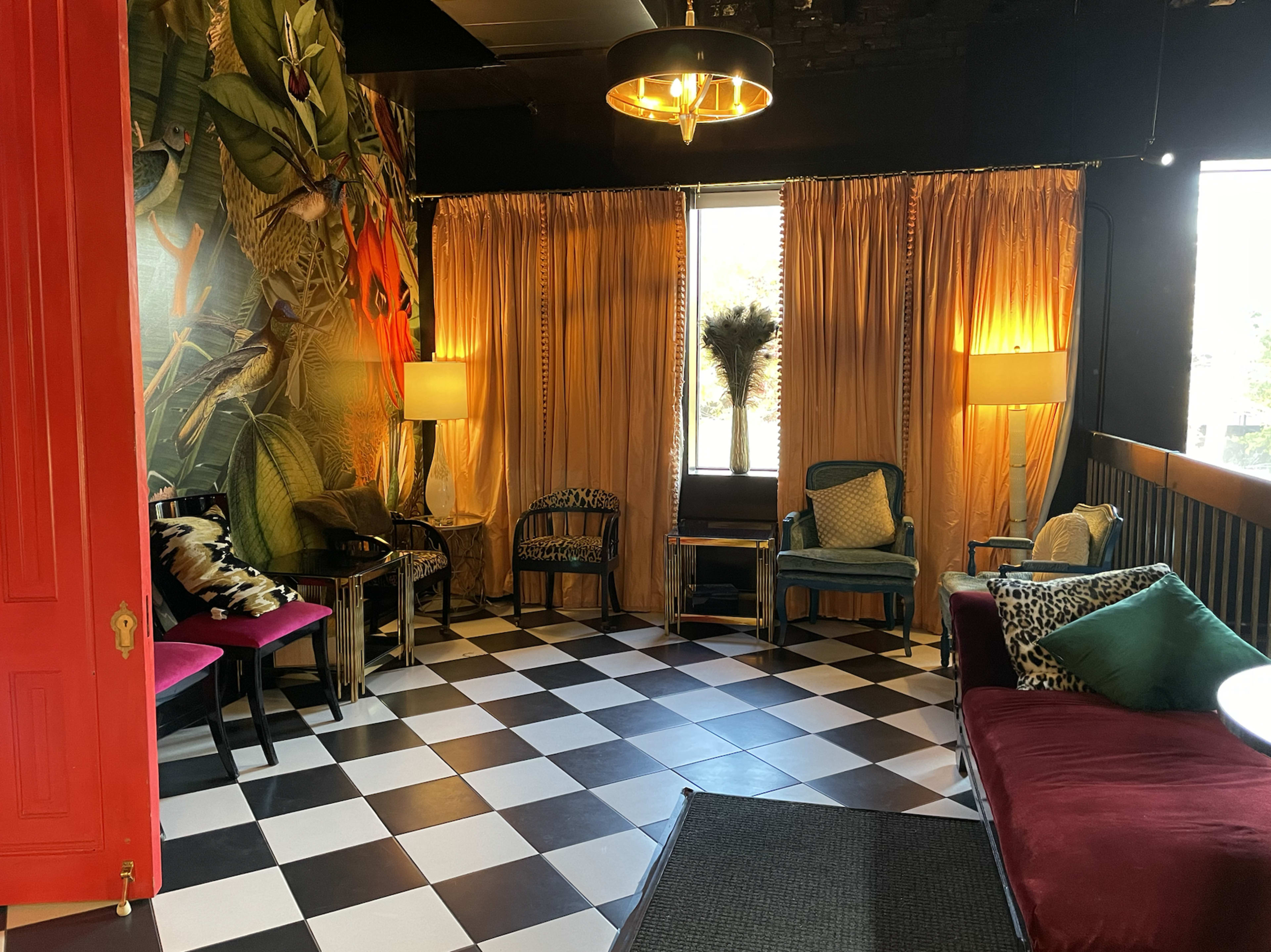 A stylish lounge area with a black and white checkered floor, colorful seating options, and large curtains illuminated by warm lighting.