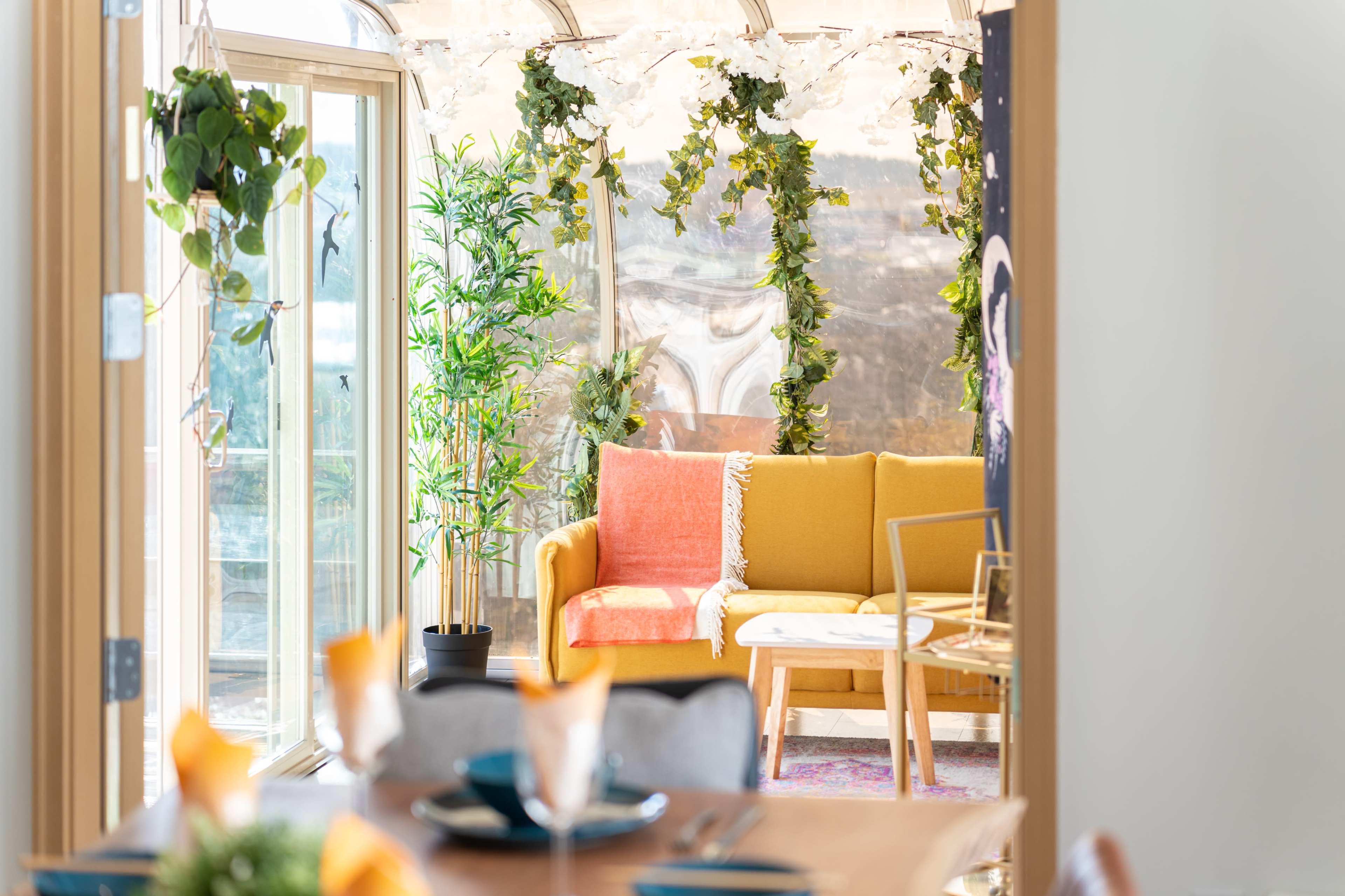 A bright, cozy living space with a yellow couch, surrounded by greenery and natural light from large windows.