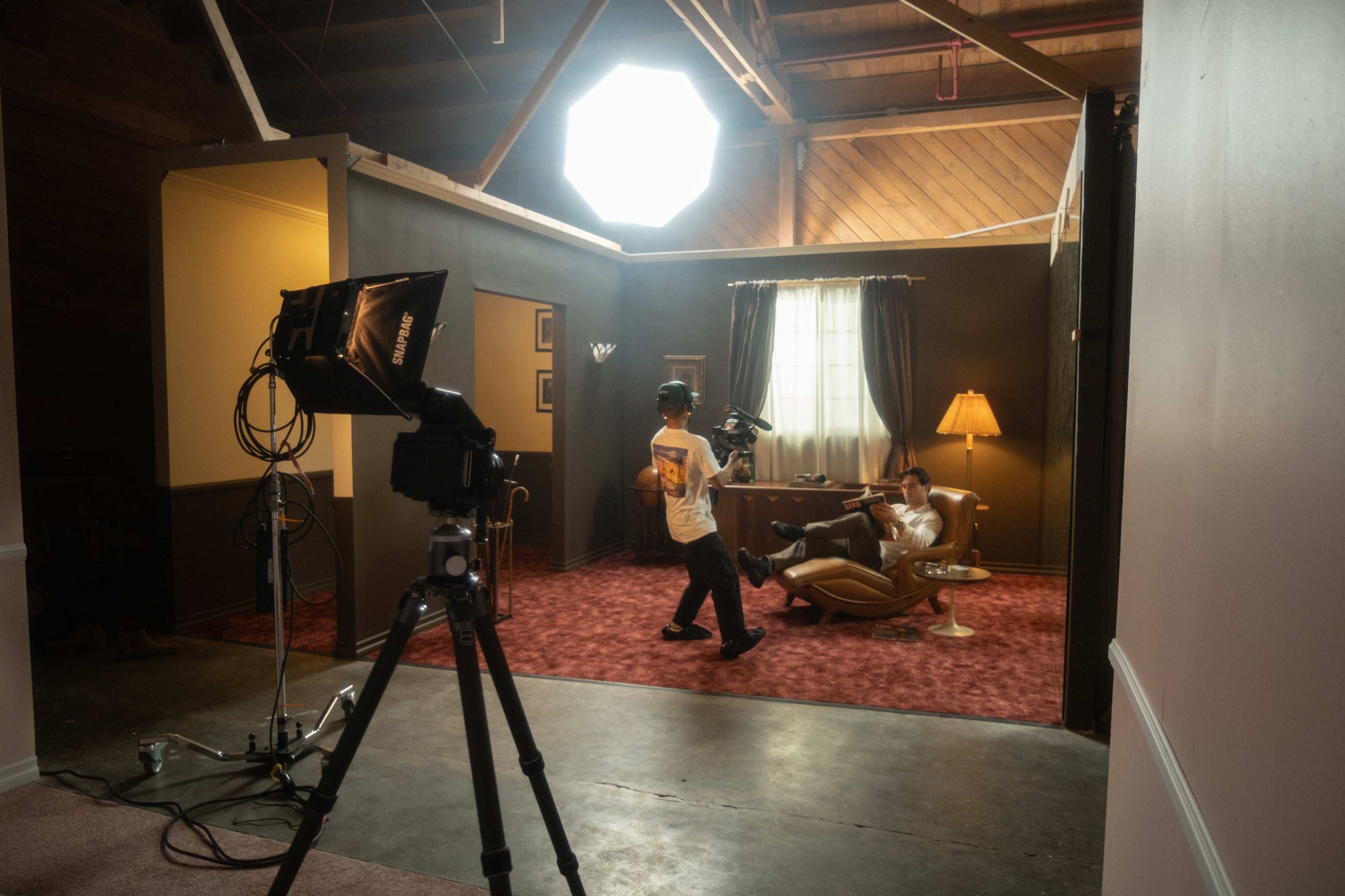A filmmaker operates a camera while a subject relaxes in an armchair, with studio lighting illuminating a vintage-style interior.