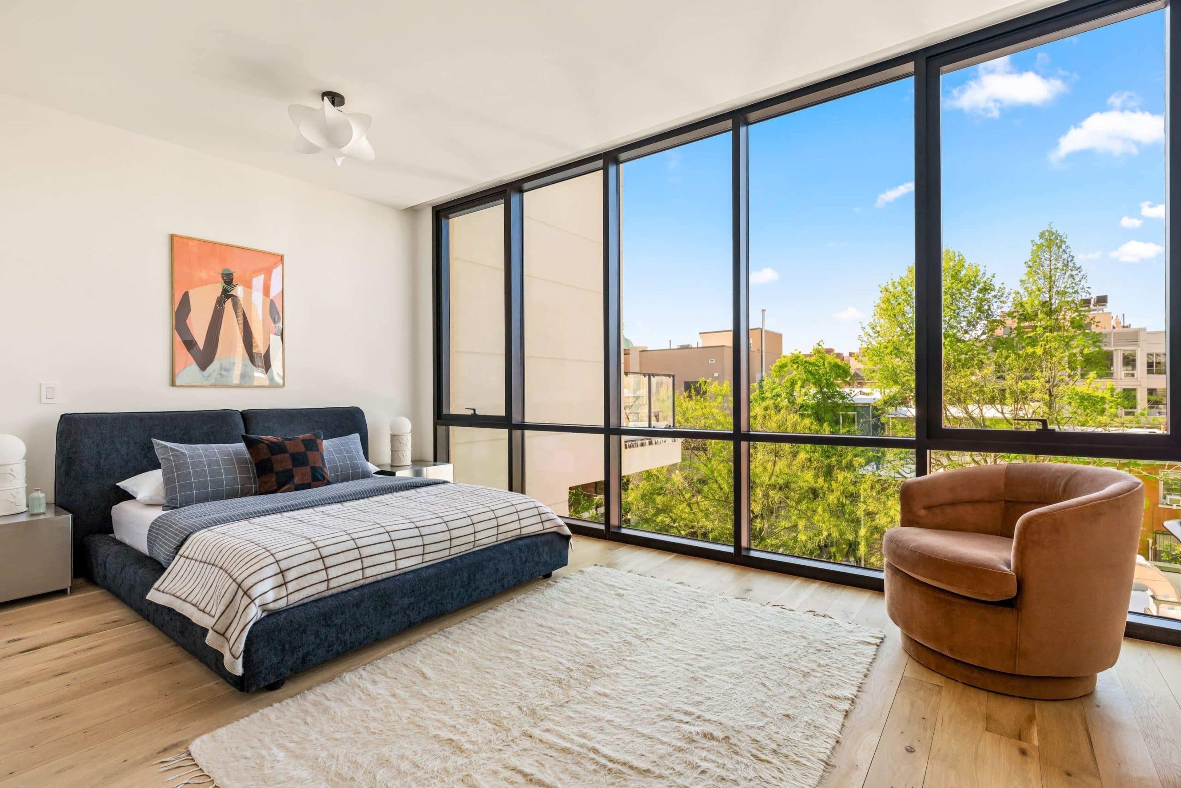 The bedroom features a bed with a navy headboard, a large window overlooking greenery, a tan armchair, and a light-colored rug on a wooden floor.