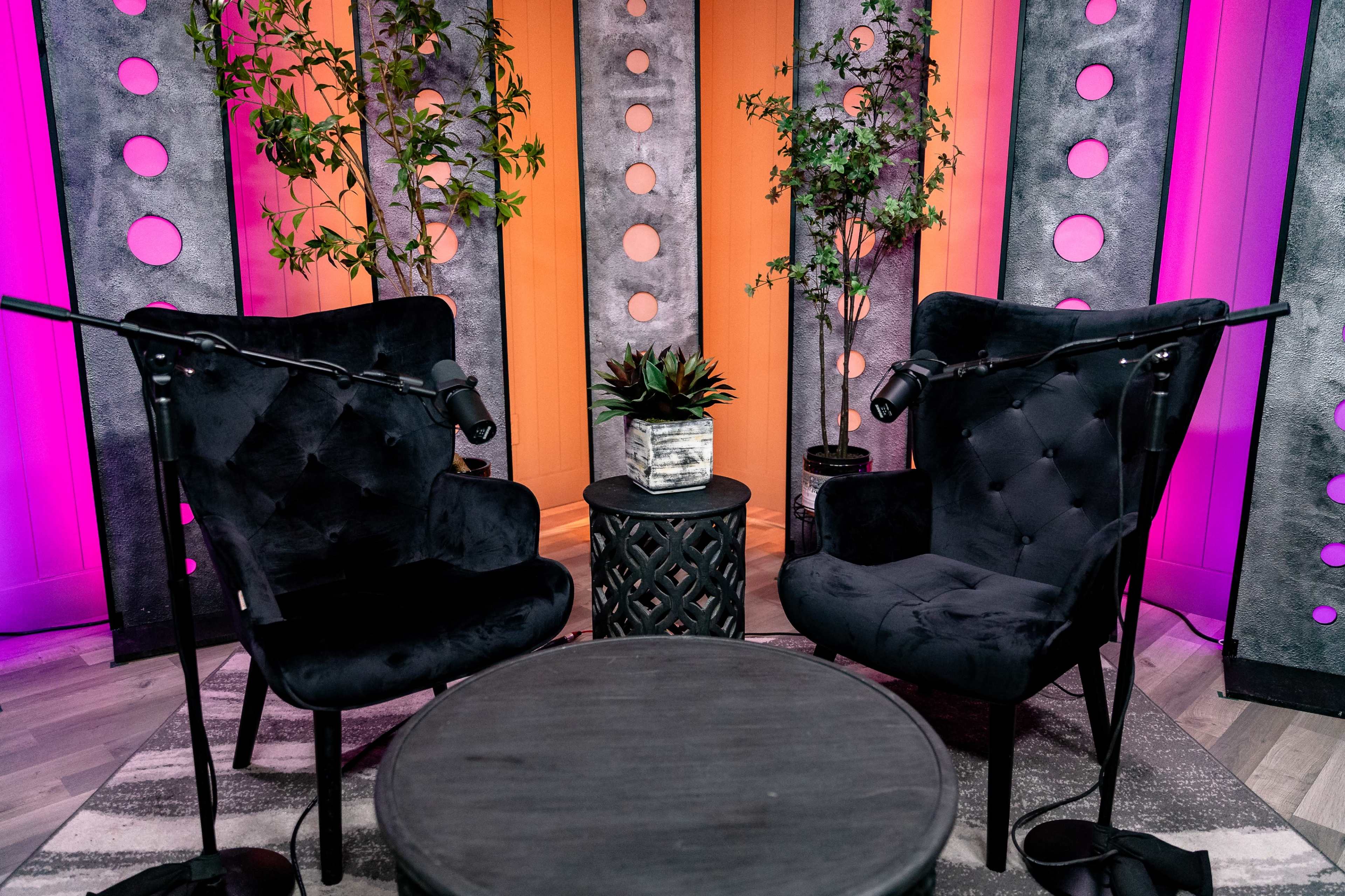 The image shows a cozy podcast or interview setup with two black chairs, a small circular table, and plants against a backdrop of brightly colored walls.