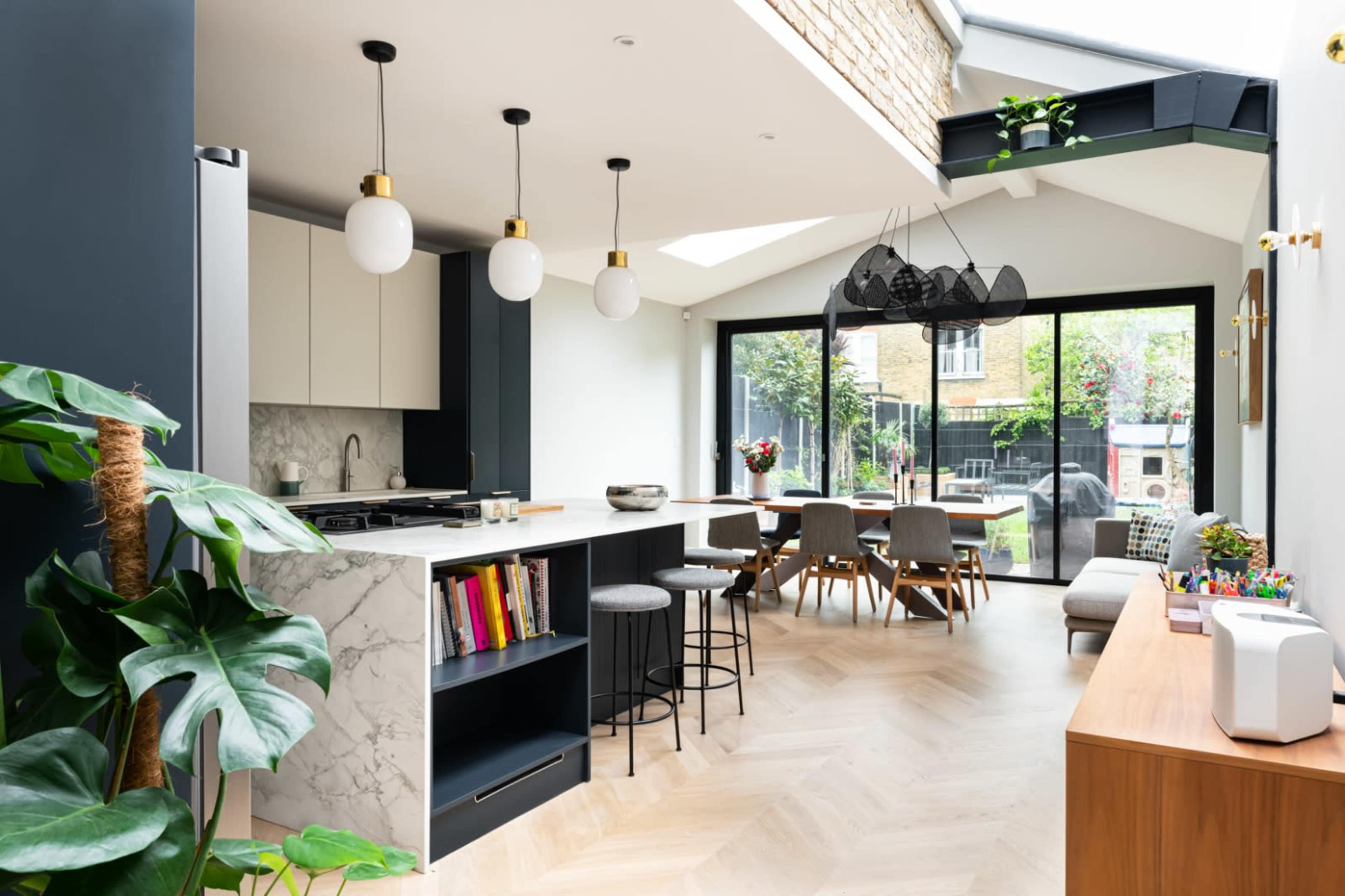 The image shows a modern kitchen and dining area featuring a marble countertop, pendant lighting, and a large window that opens to a garden.