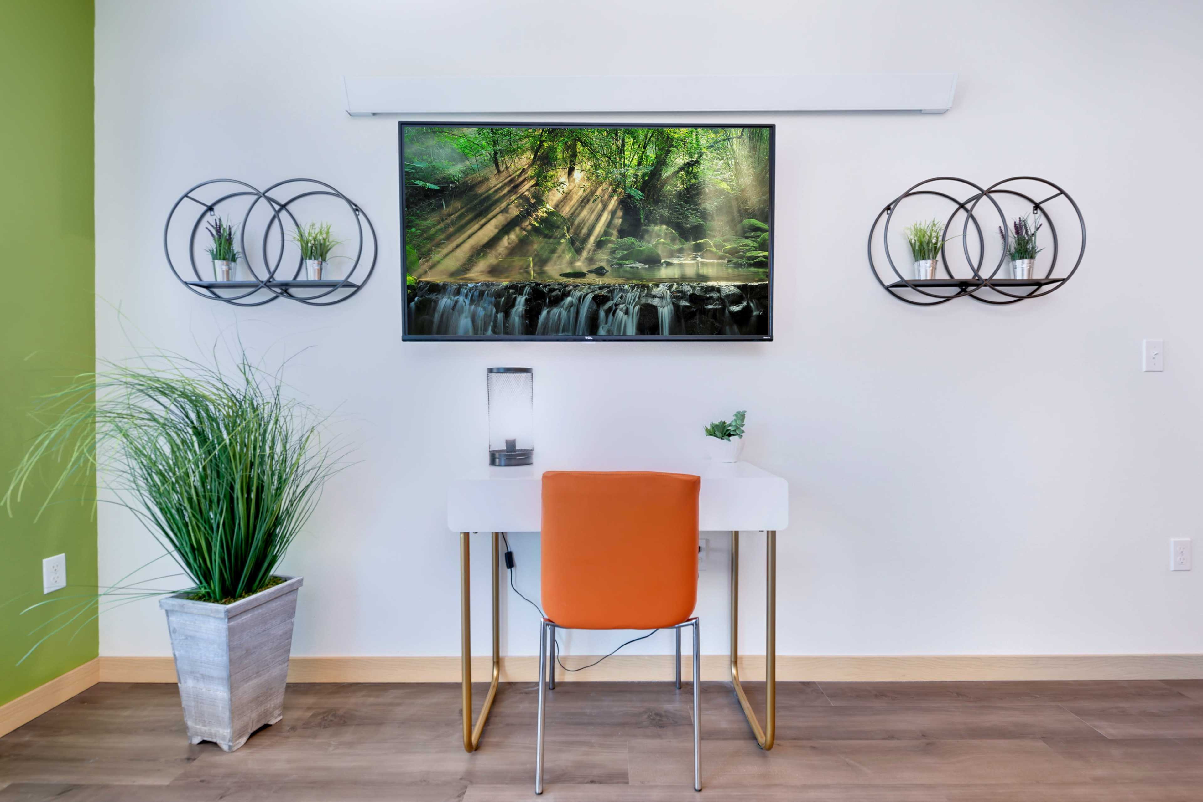 A modern workspace features a white desk and an orange chair, with a wall-mounted screen displaying a nature scene, flanked by circular shelves holding small plants.