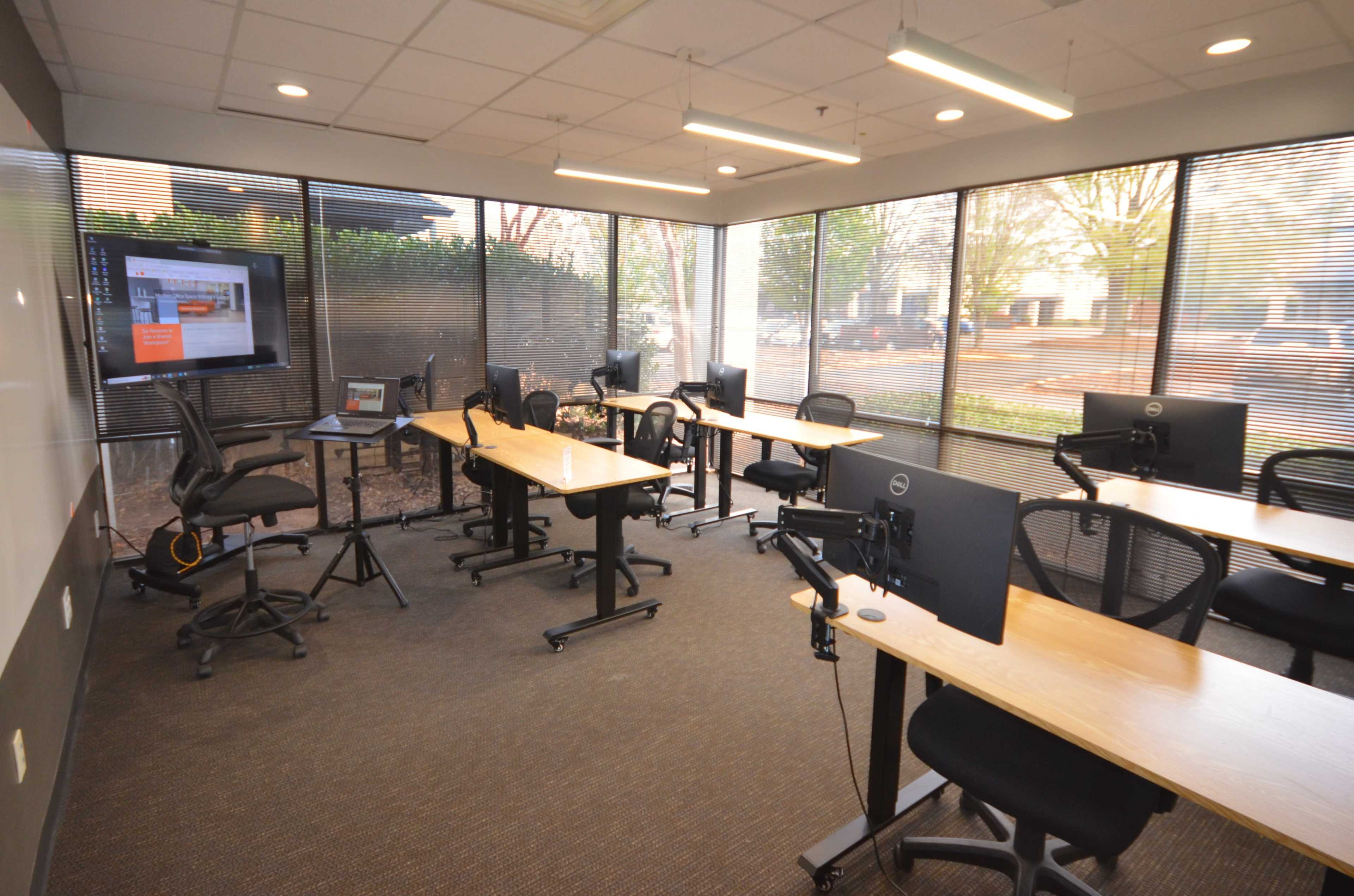 The image shows a modern classroom with several desks and computer monitors, surrounded by large windows that provide natural light.