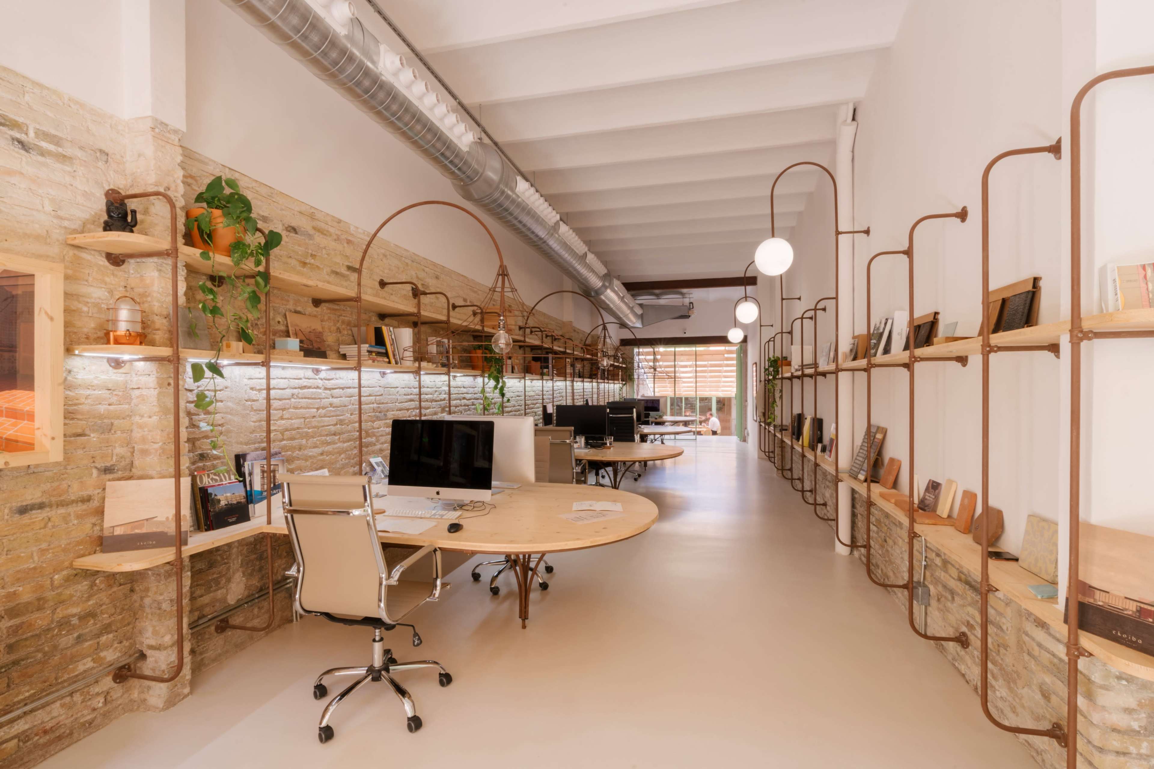 The image shows a modern open office space with wooden shelves, computers on desks, and plants along the walls, illuminated by pendant lights.
