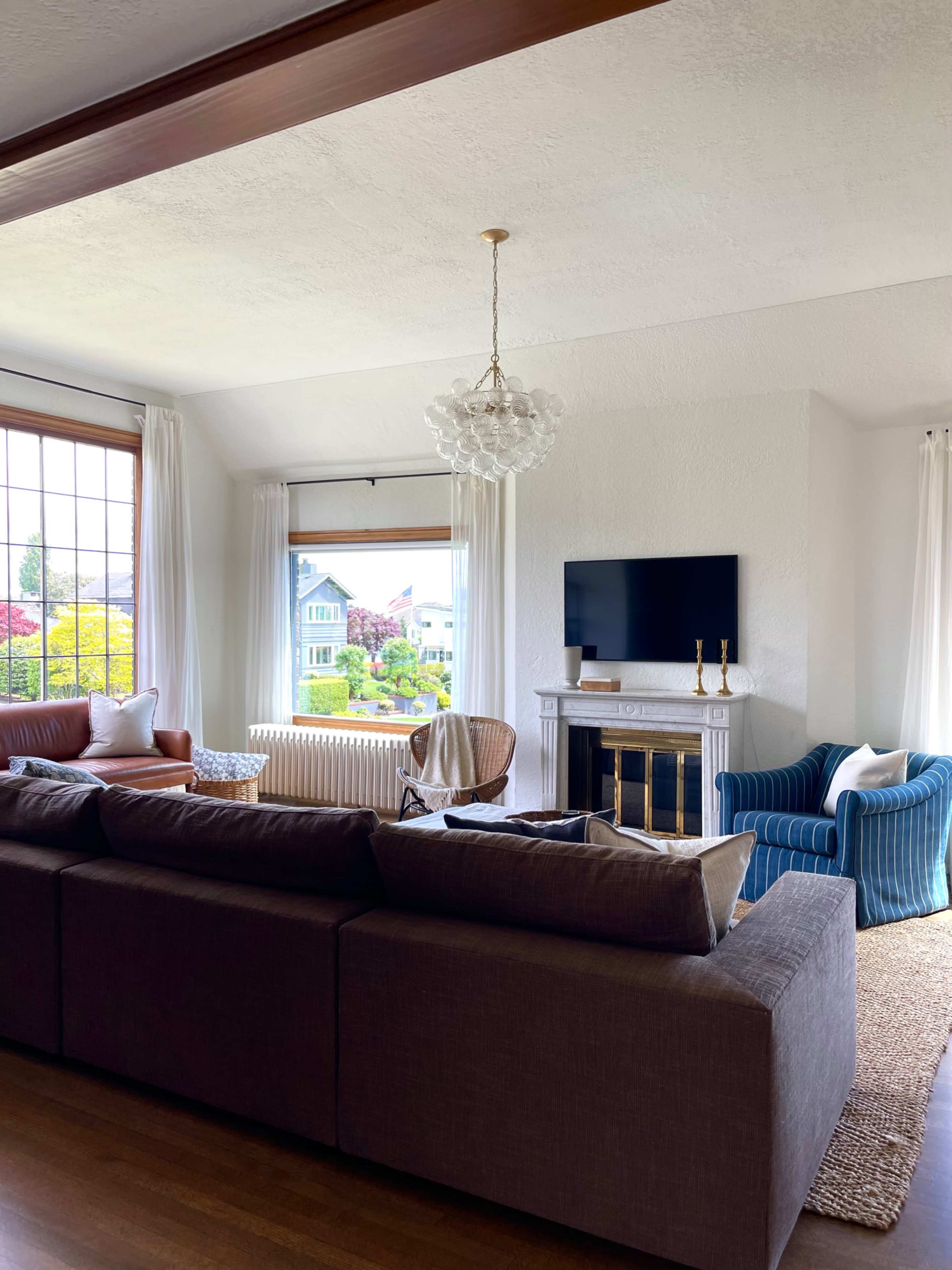 A cozy living room features a large sectional sofa, a fireplace with a television mounted above it, and windows that offer a view of an outdoor garden.