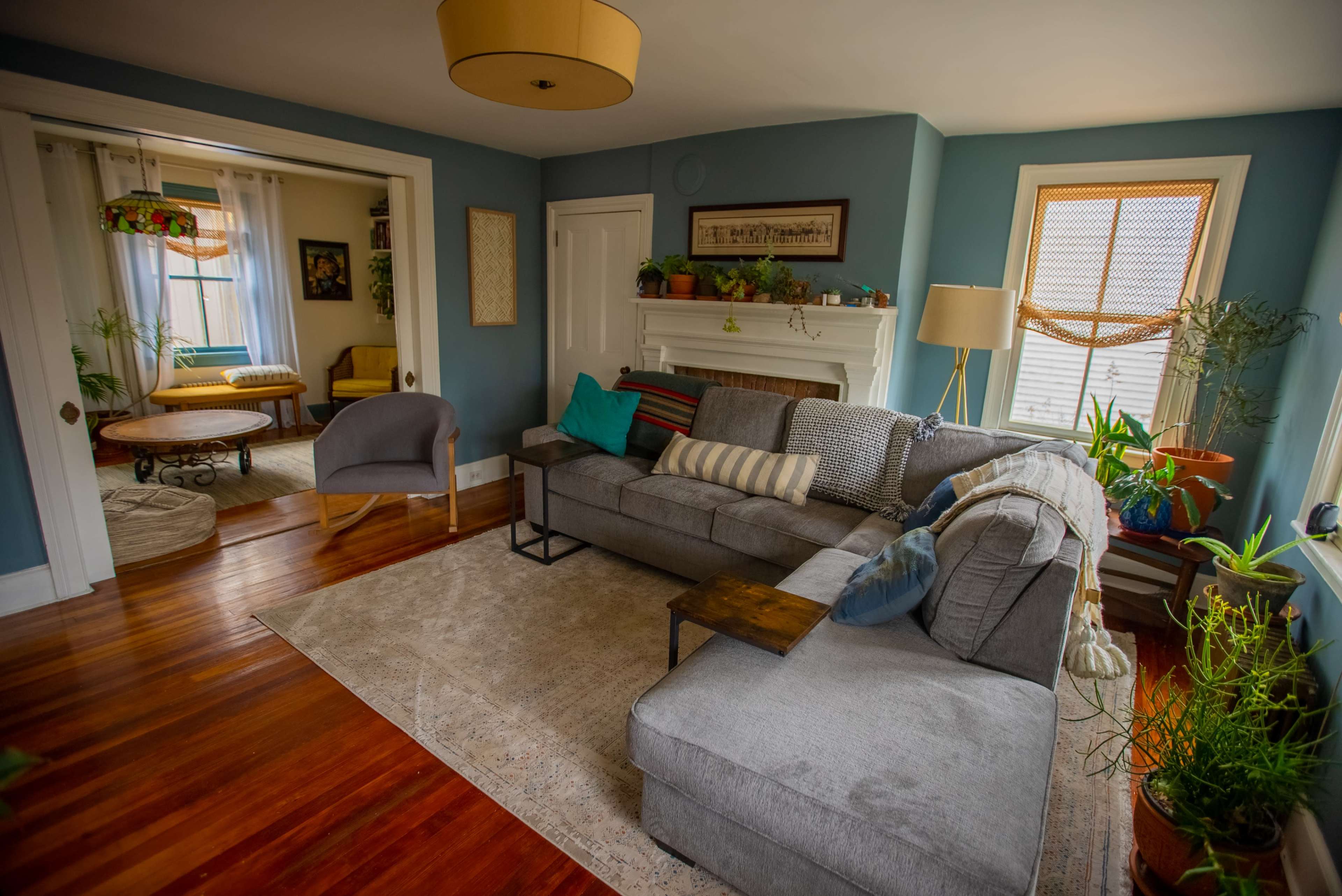 The image shows a cozy living room featuring a sectional sofa, a wooden side table, and several plants, with a doorway leading to another room in the background.