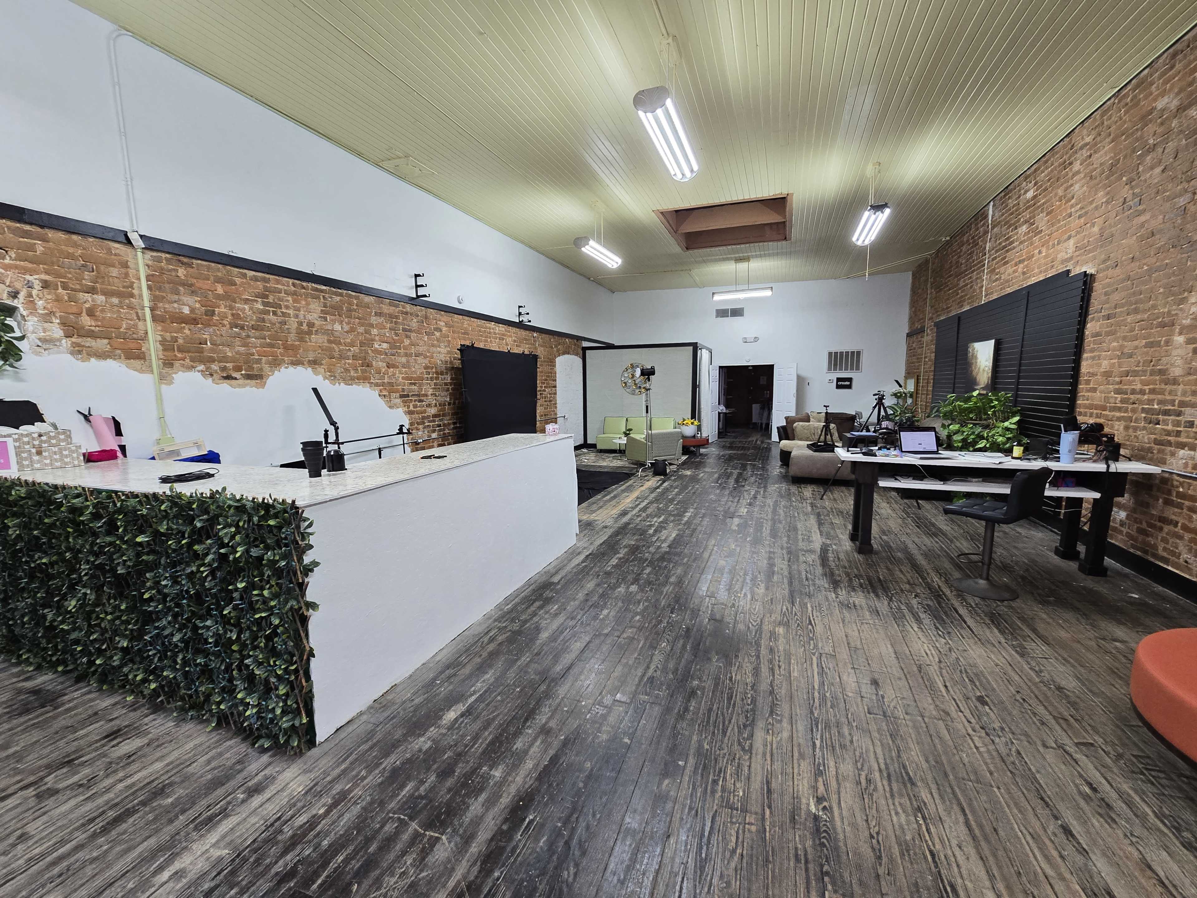 The image shows an office space with exposed brick walls, a reception area with a white counter, and a workspace featuring multiple desks and chairs.