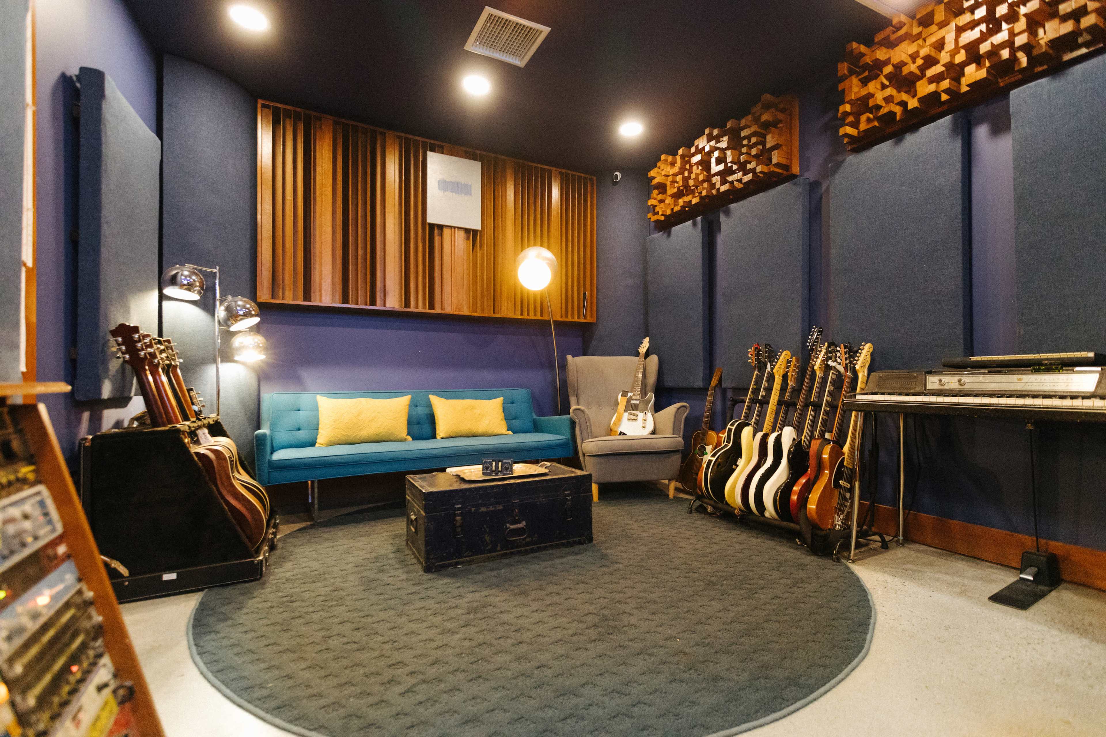 A music studio features a blue couch, a gray armchair, a round rug, a collection of guitars, and soundproofing panels on the walls.