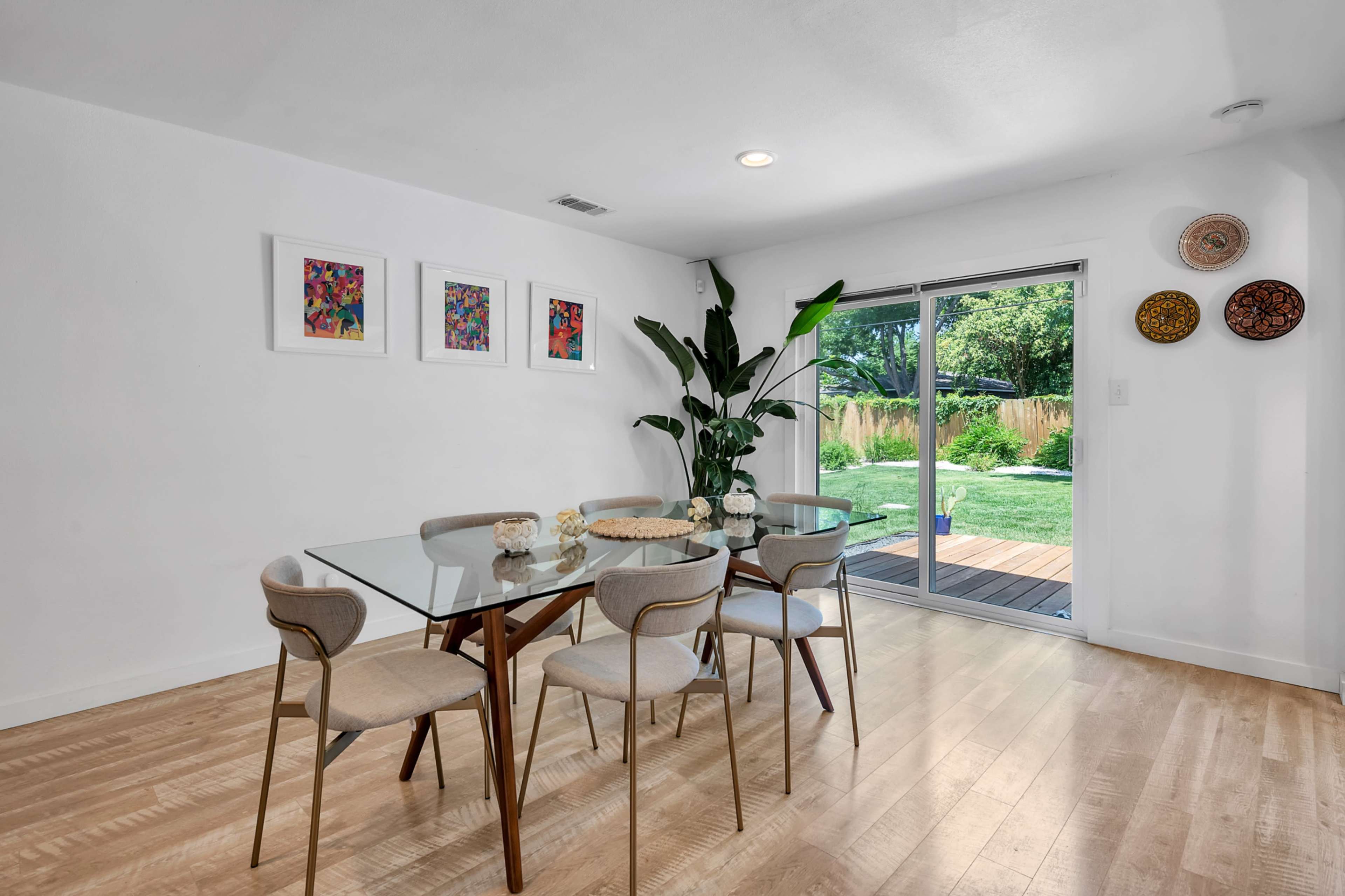 A modern dining area features a glass table surrounded by six chairs, with artwork on the walls and a sliding glass door leading to a garden.
