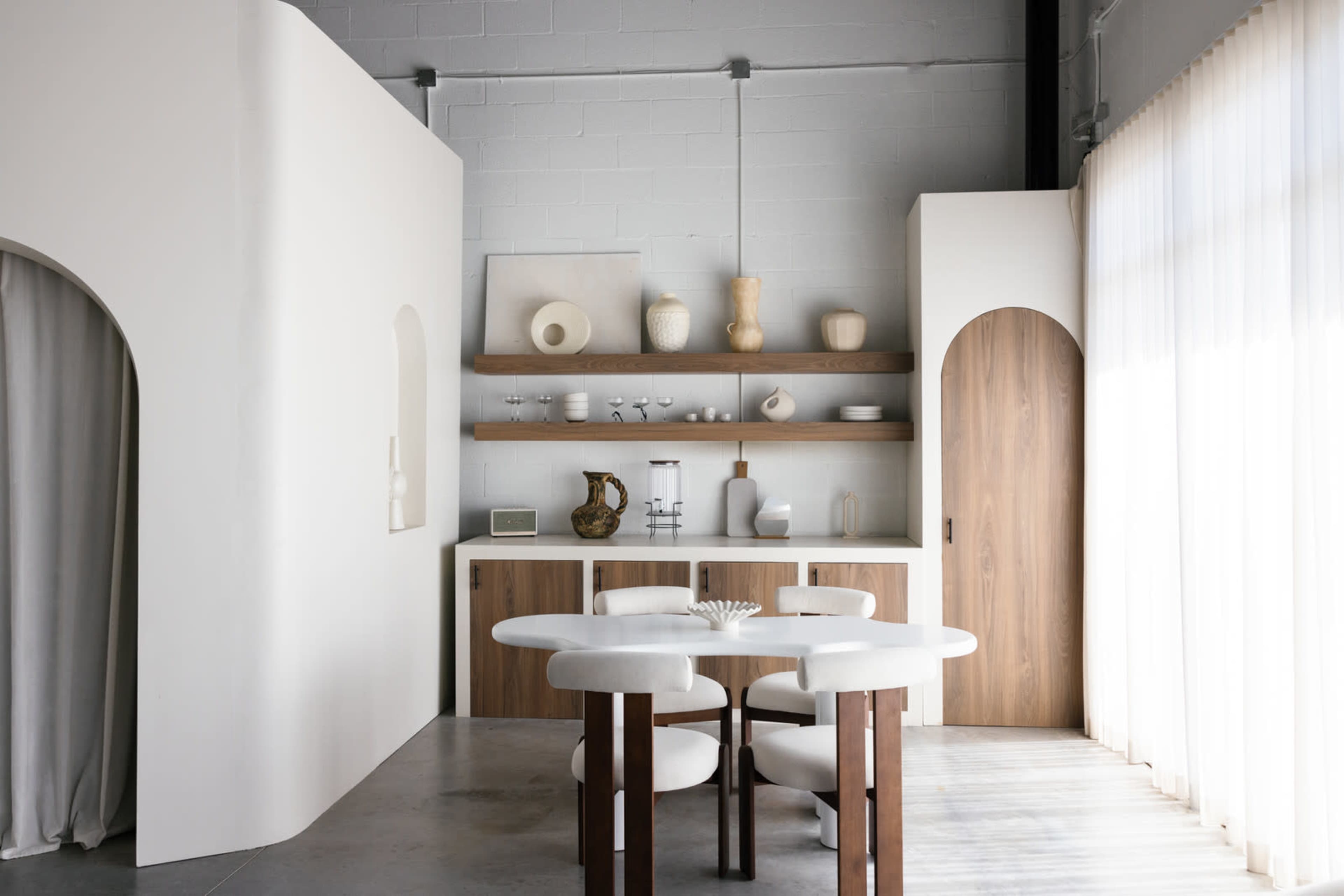A minimalist dining area features a round table surrounded by four chairs, against a backdrop of light-colored walls and wooden shelves displaying various decor items.