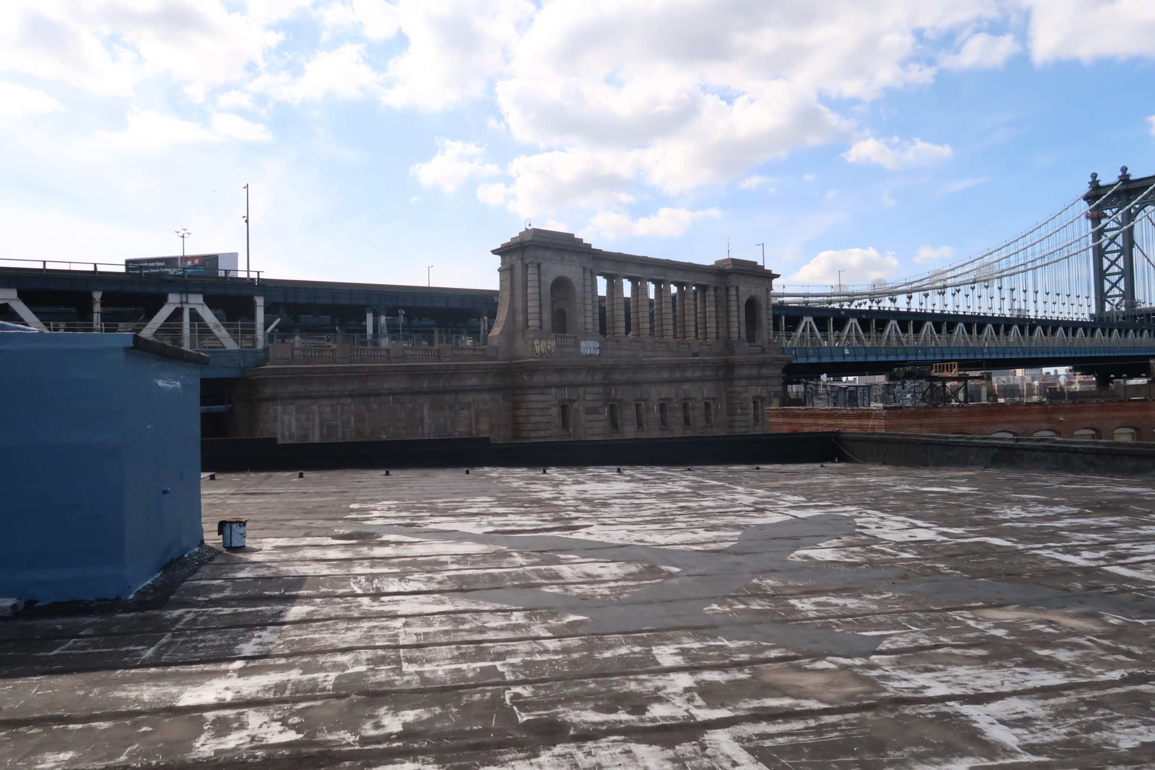 A weathered rooftop with a bridge and a historic stone structure in the background, set against a cloudy sky.