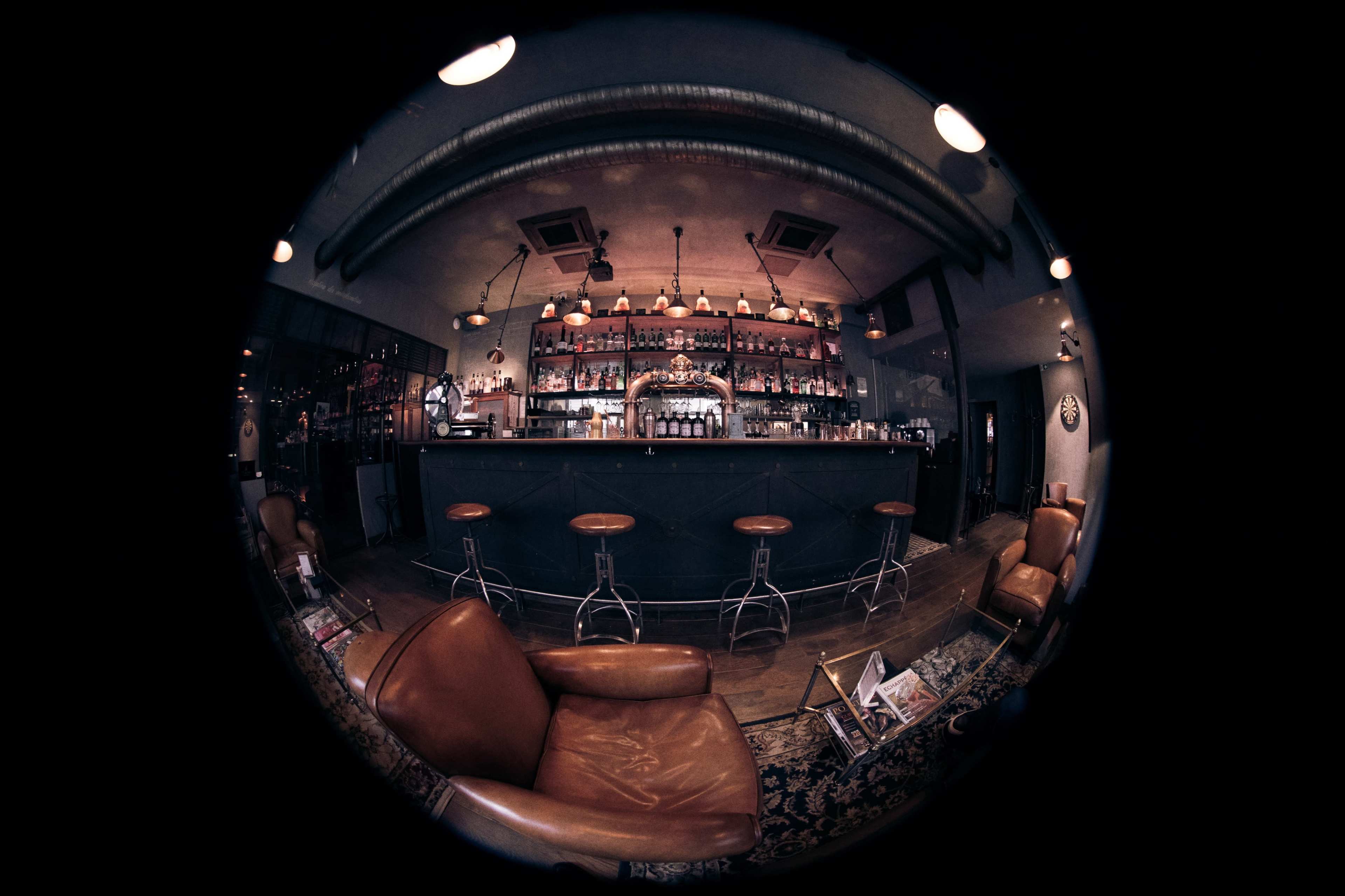 A vintage-style bar with a long counter, high stools, and shelves filled with bottles is depicted in a cozy interior setting.