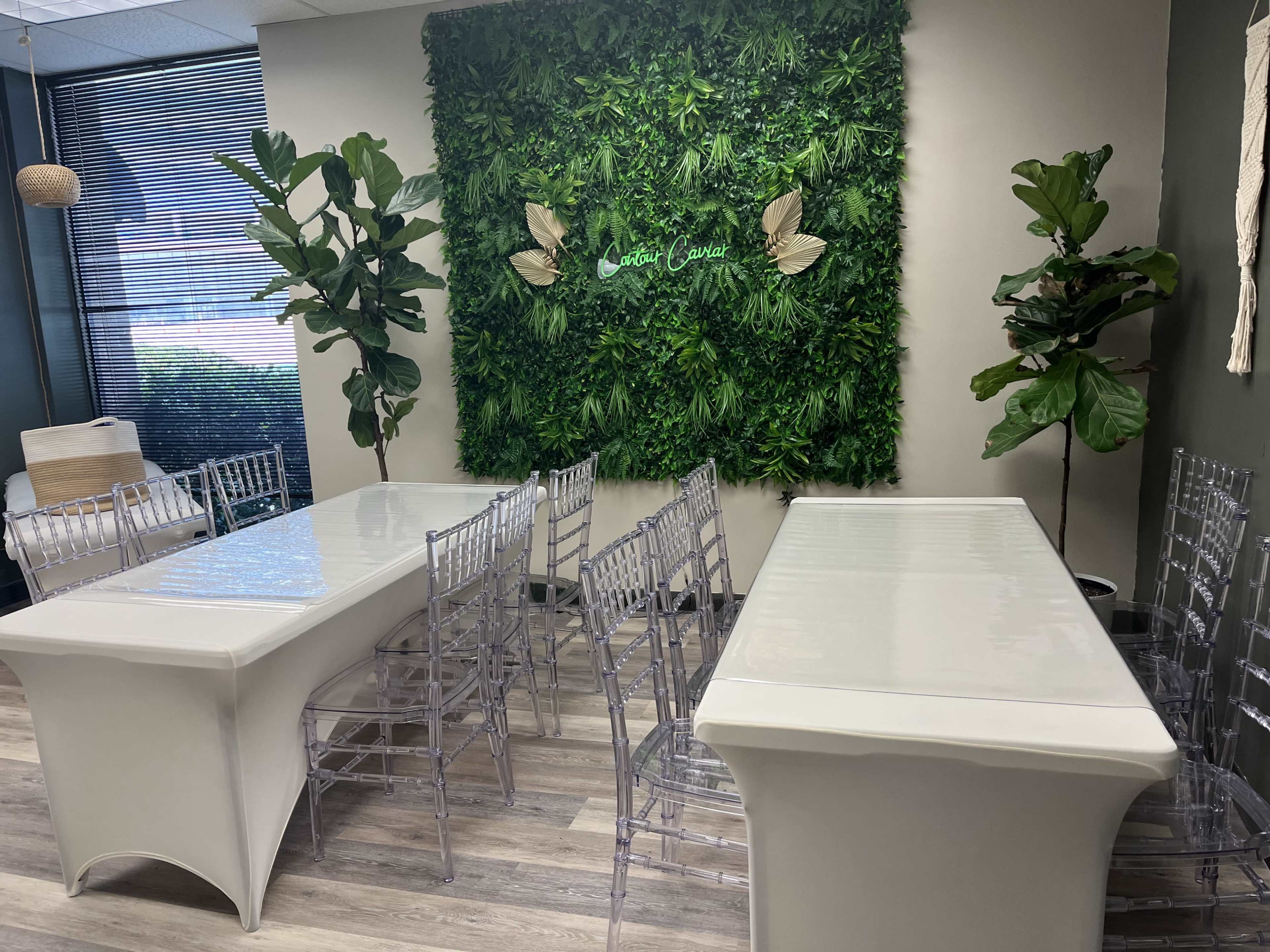 A modern room with two white tables, clear plastic chairs, and a green wall adorned with plants and decorative elements.