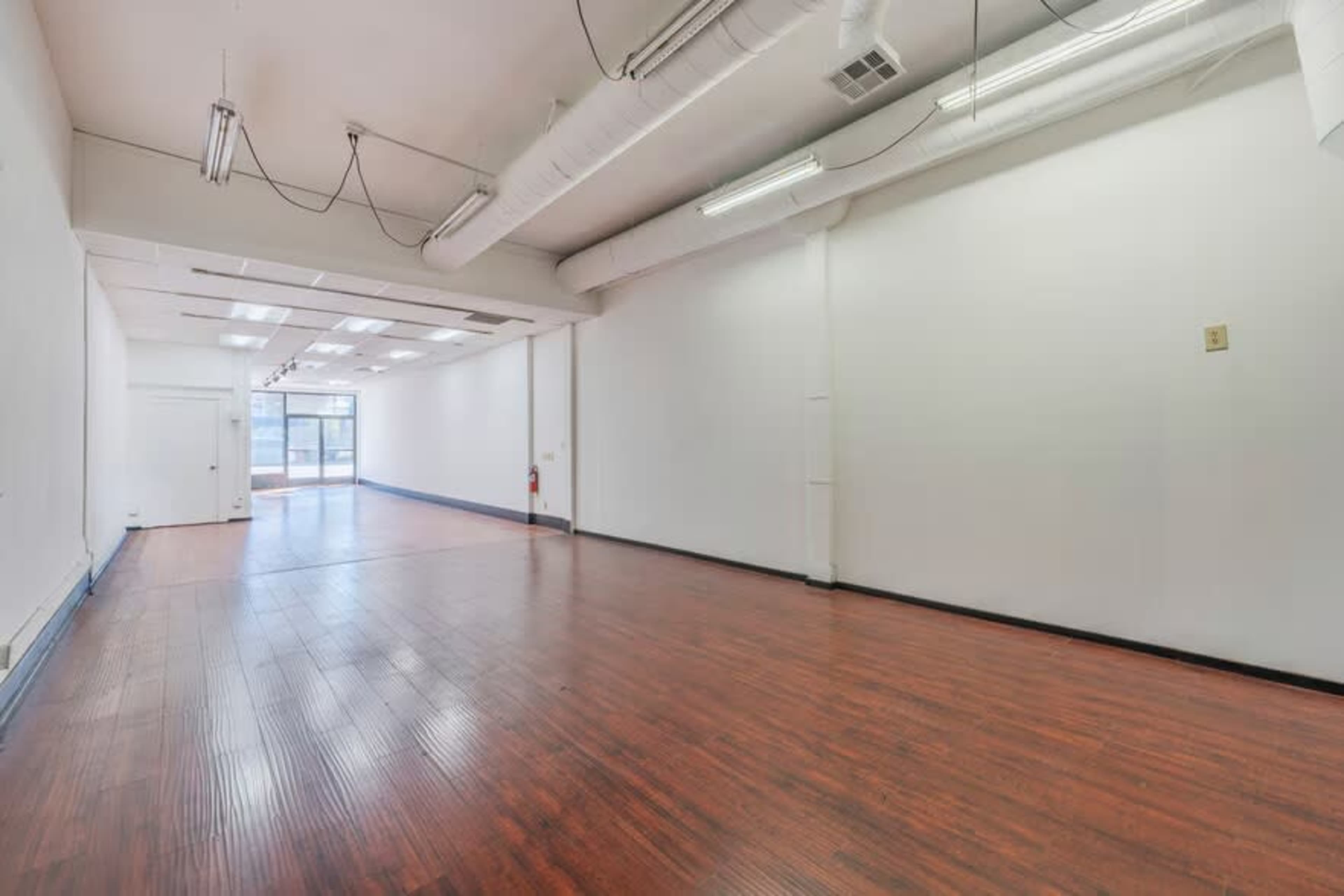 The image shows a spacious, empty room with wooden flooring and white walls, featuring overhead lighting and a door leading to another area.