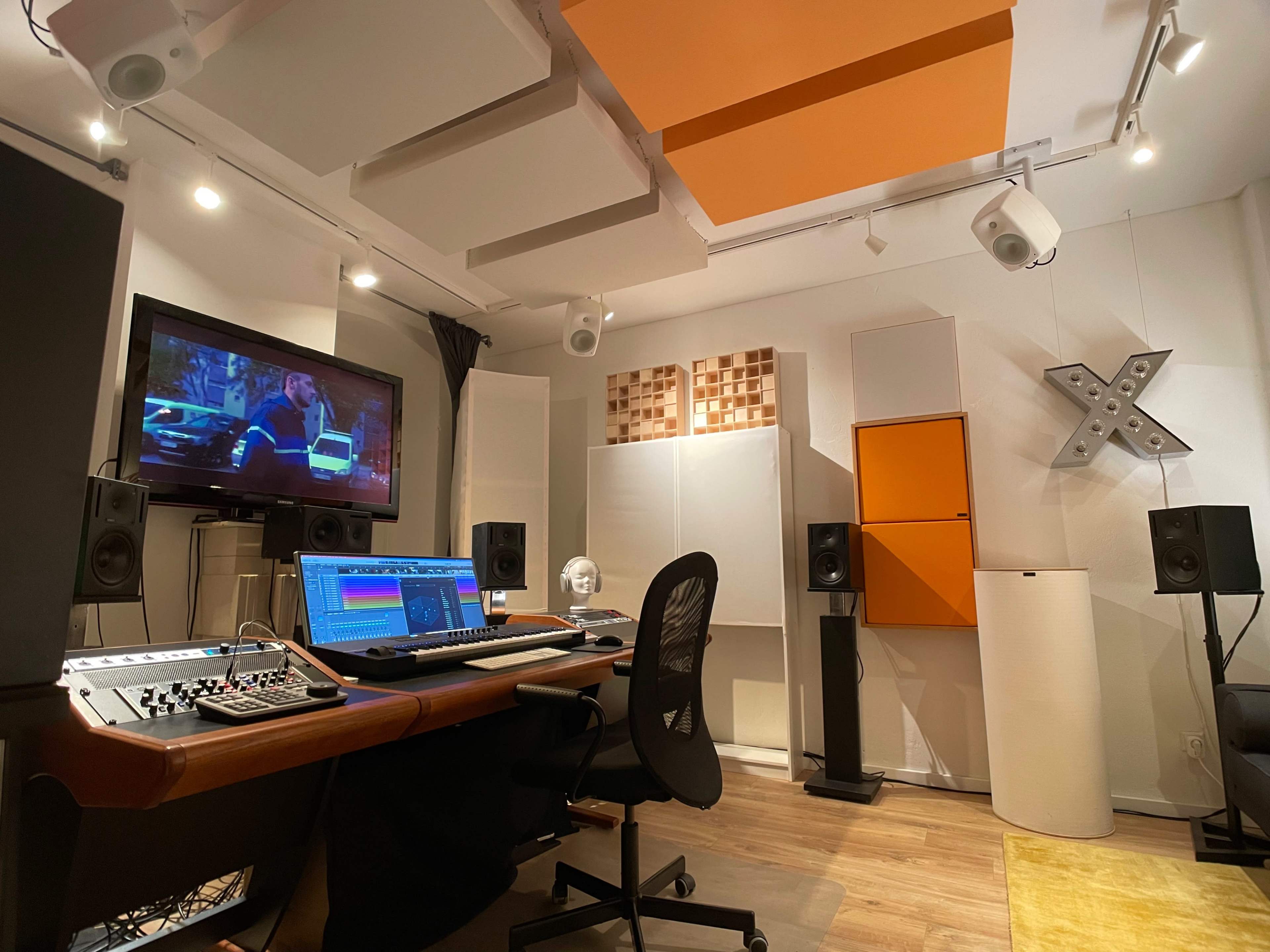 A music production studio equipped with a mixing console, computer, speakers, and soundproofing panels, featuring a television display showing a scene from a film.