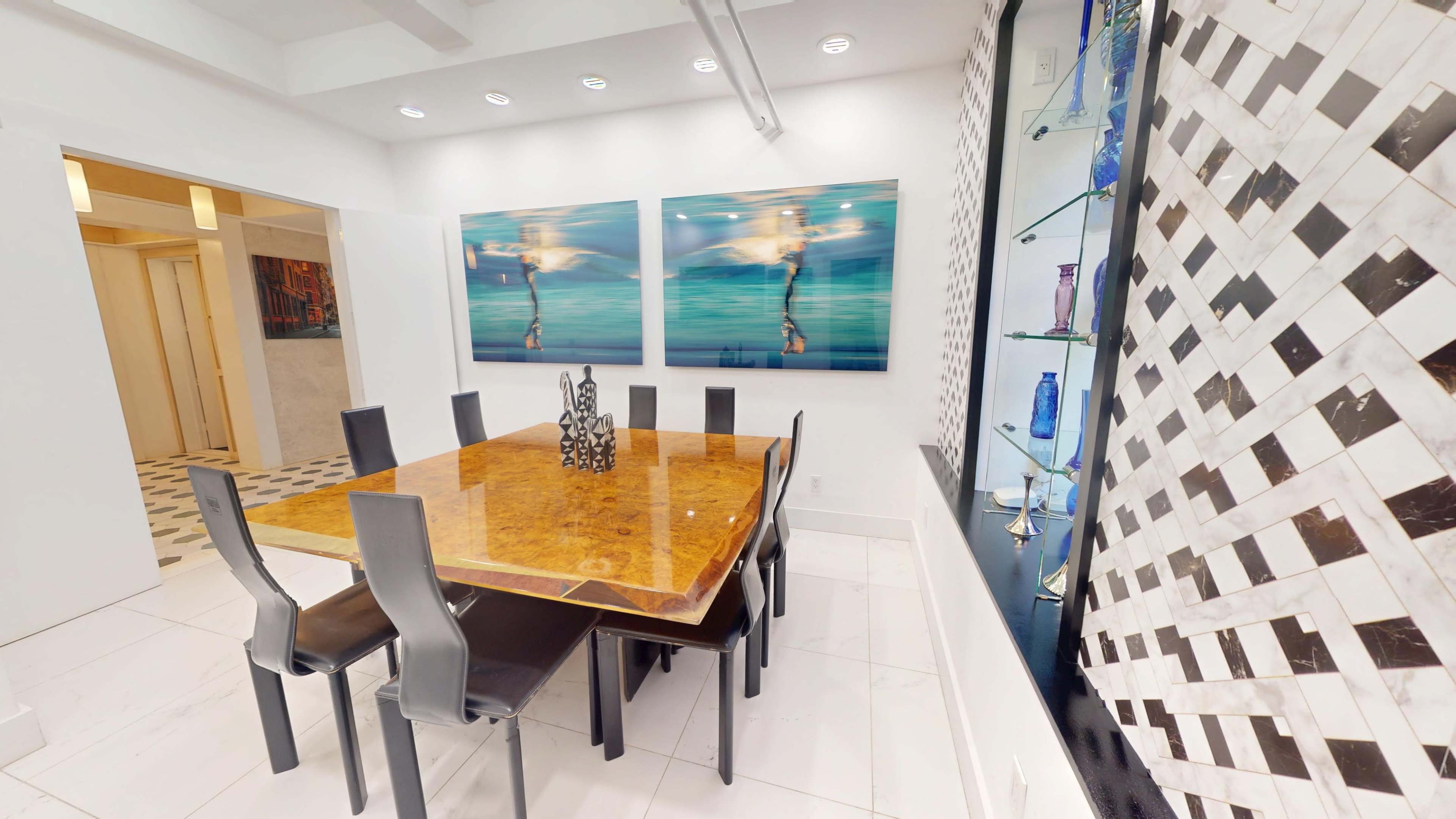 A modern dining area features a large wooden table surrounded by black chairs, with two art pieces on the wall and a display shelf showcasing decorative items.