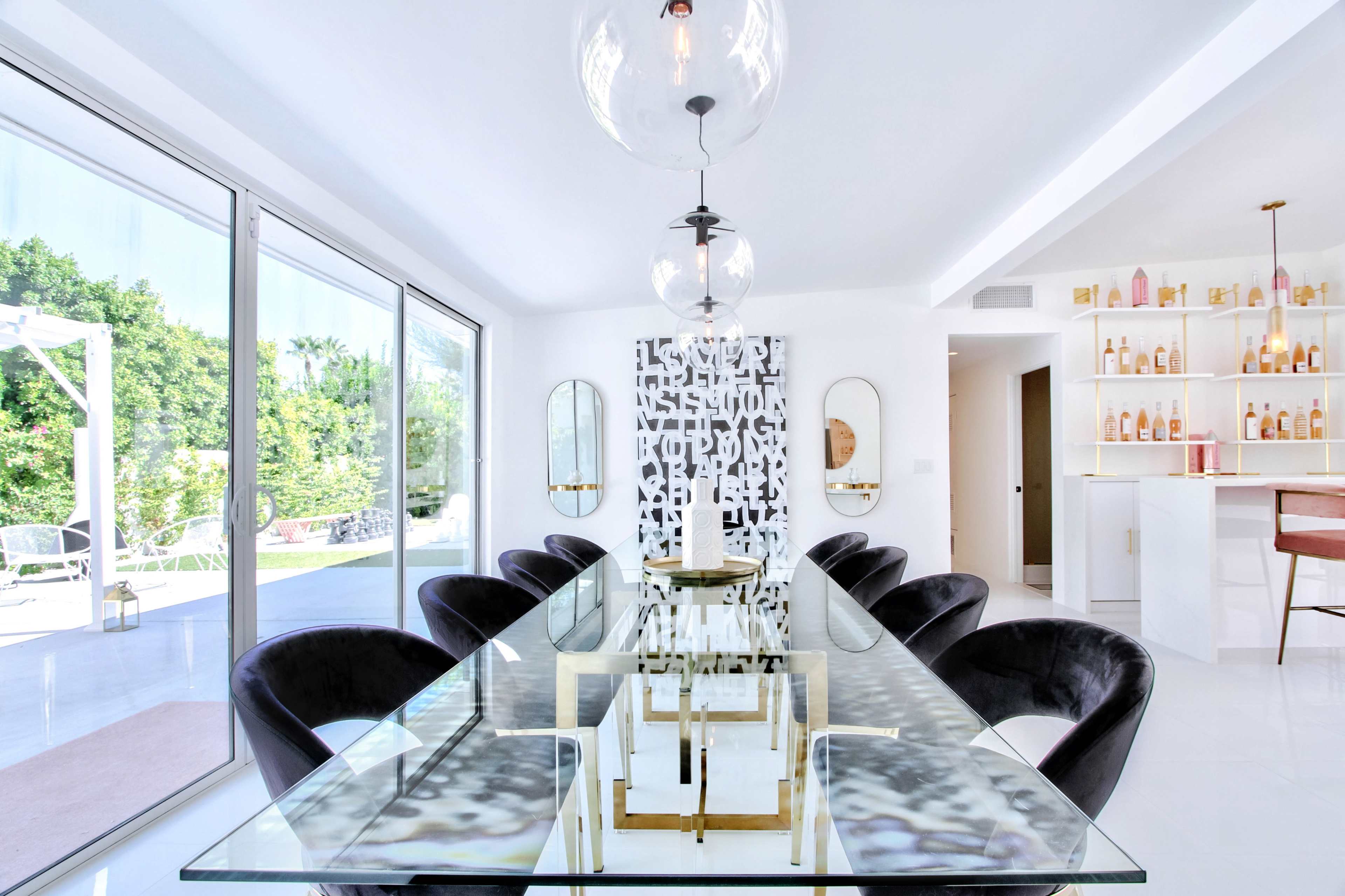 A modern dining room features a glass table surrounded by black chairs, with large windows overlooking a patio and greenery.