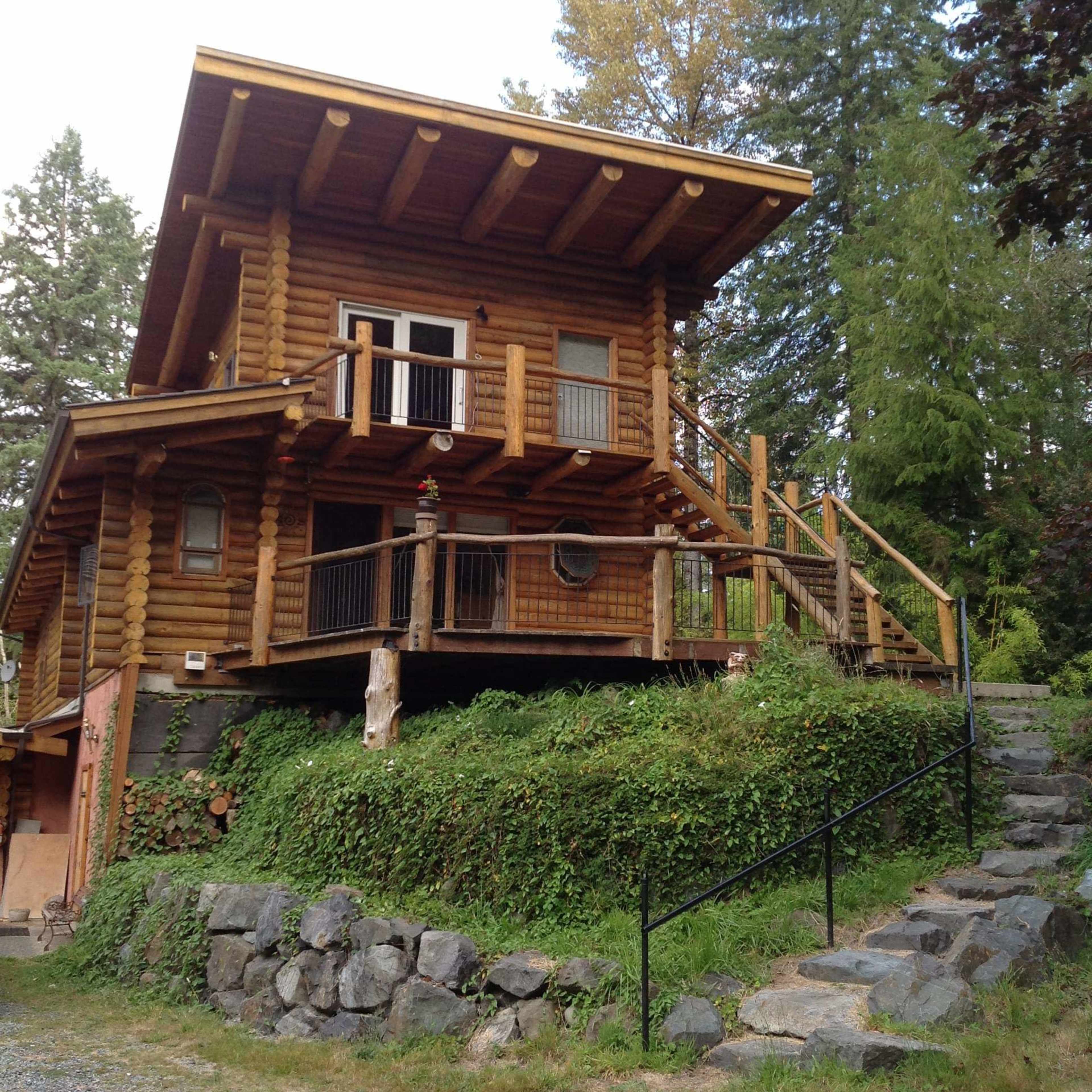 A two-story log cabin with a wooden deck is situated on a grassy slope surrounded by trees and a stone pathway.