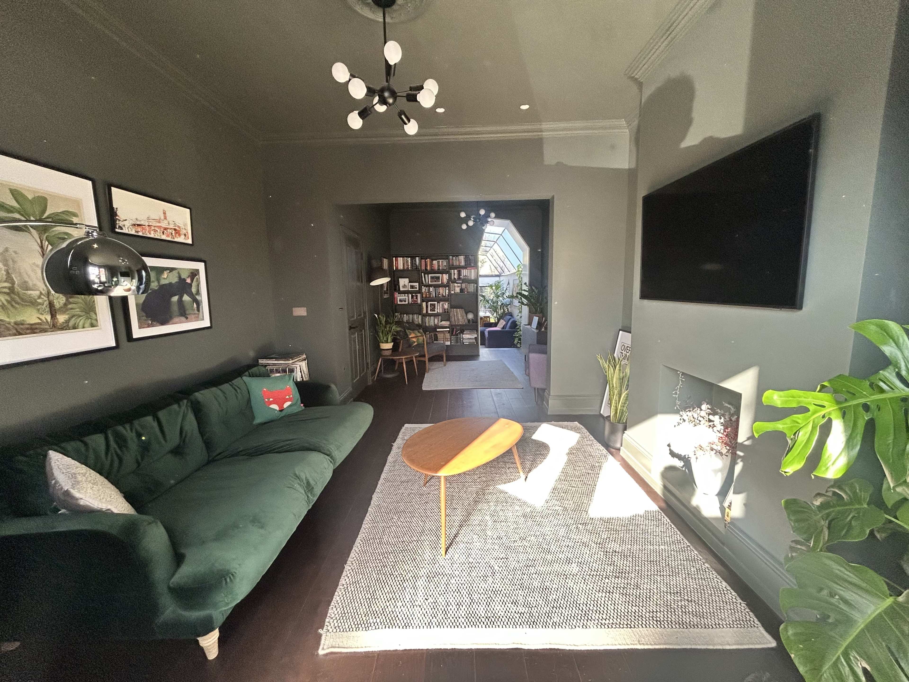 The image shows a stylish living room with dark green walls, a green sofa, and a light wood coffee table on a textured rug.