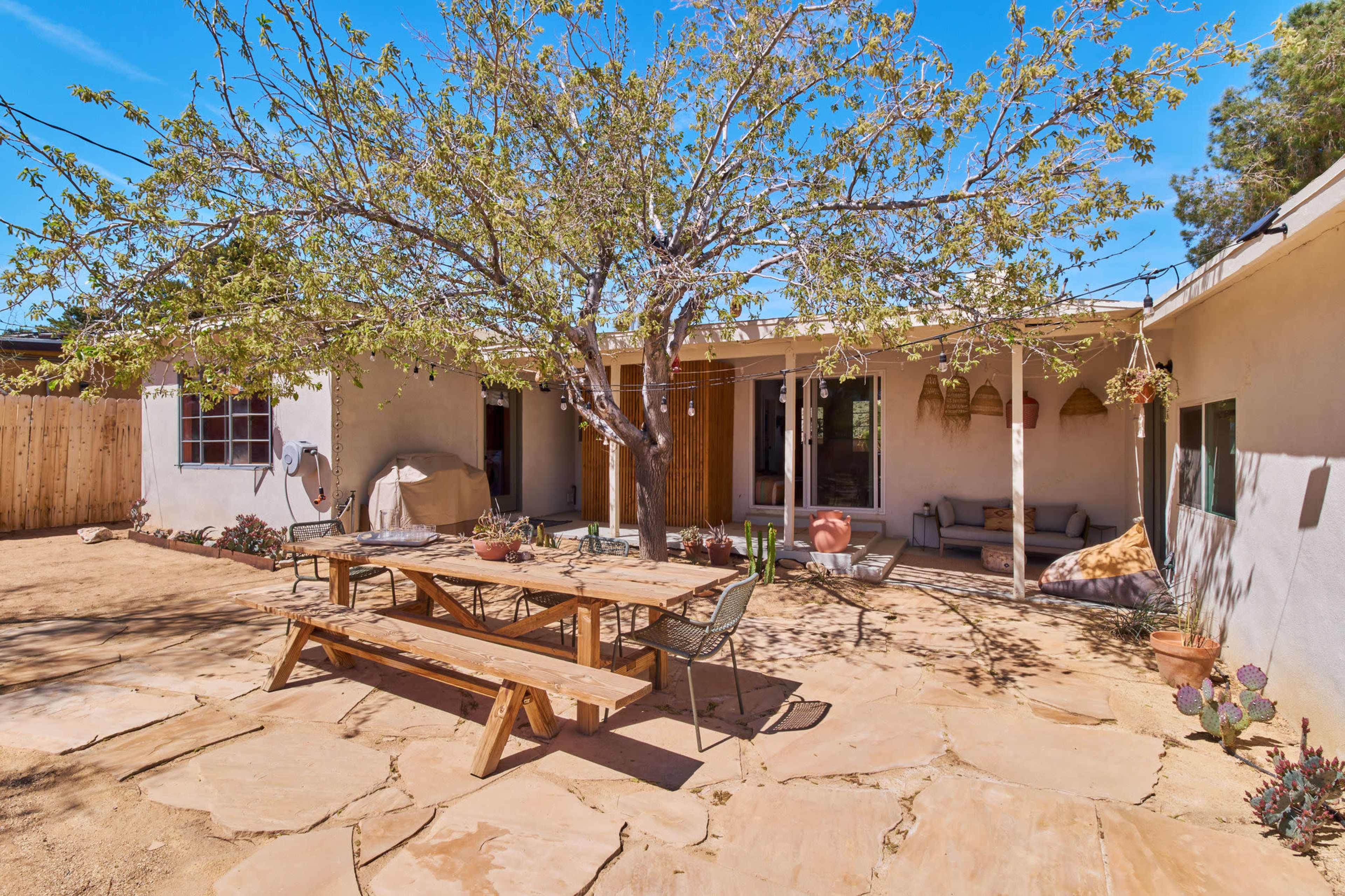 A spacious courtyard features a large wooden picnic table surrounded by chairs, with a tree providing shade in the center.