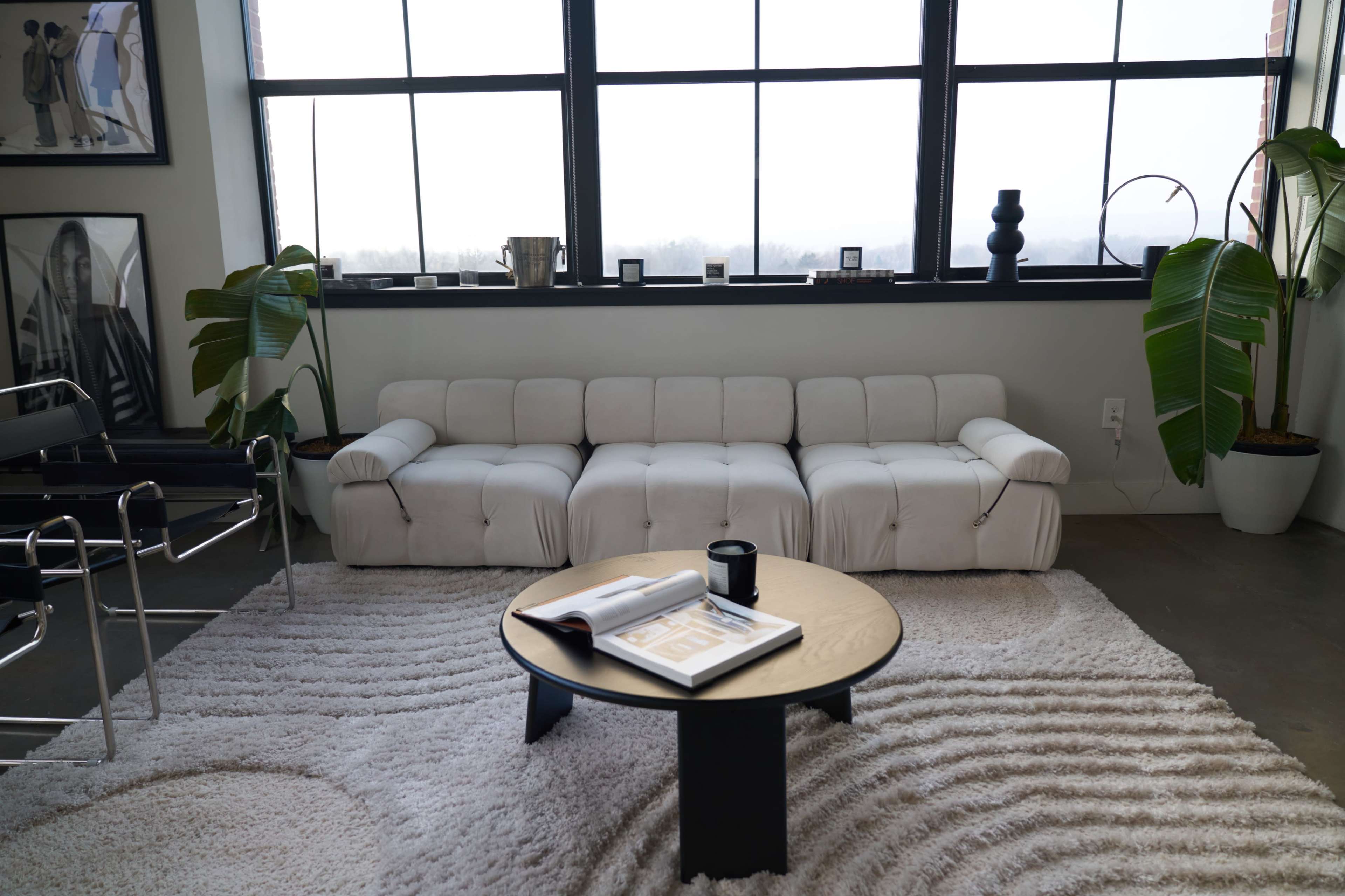 A white sectional sofa is positioned in front of large windows, with a round coffee table and a patterned rug nearby.