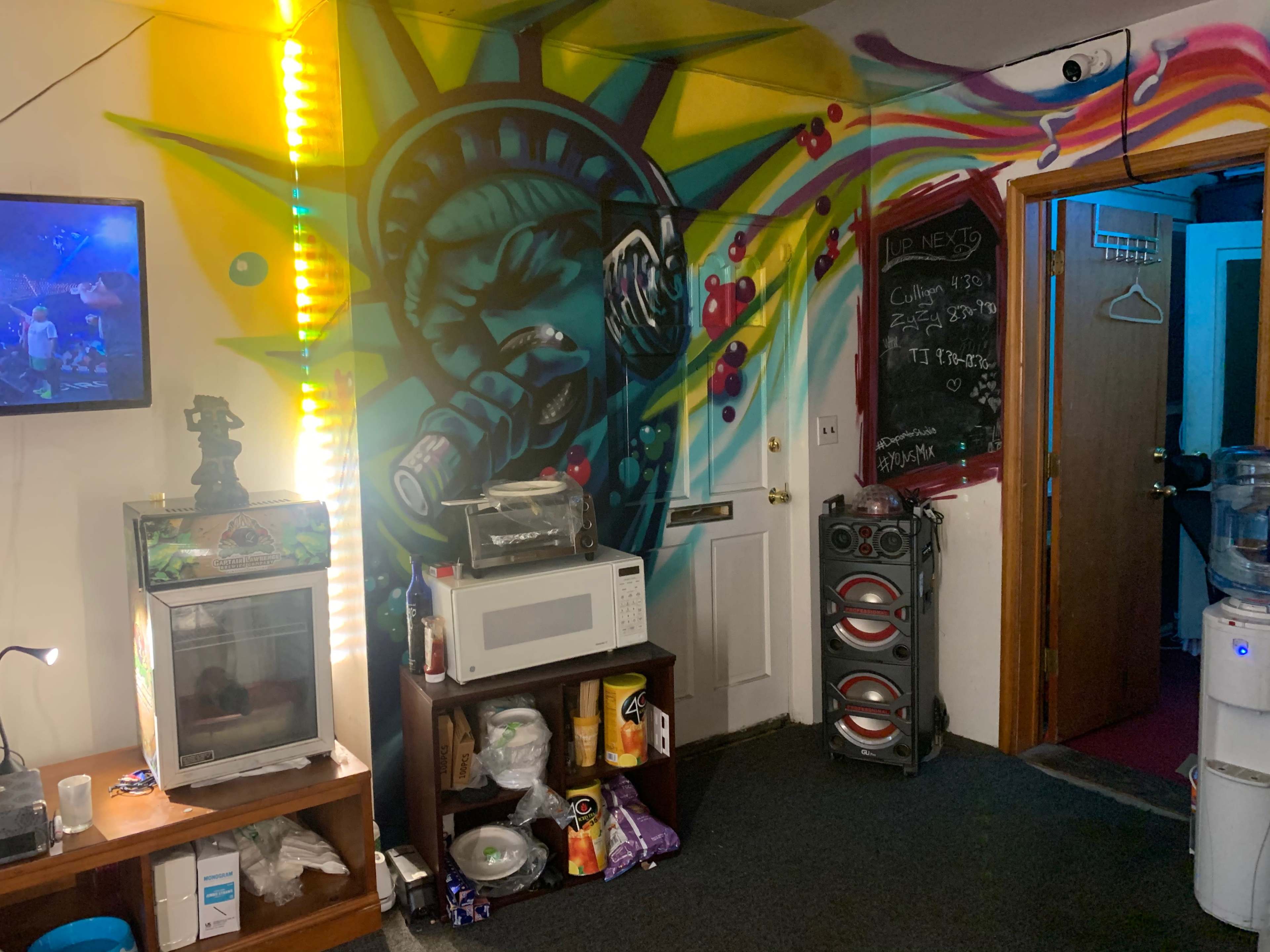 A room featuring a colorful mural of the Statue of Liberty, a microwave, a mini fridge, and a speaker system, with a television mounted on the wall.