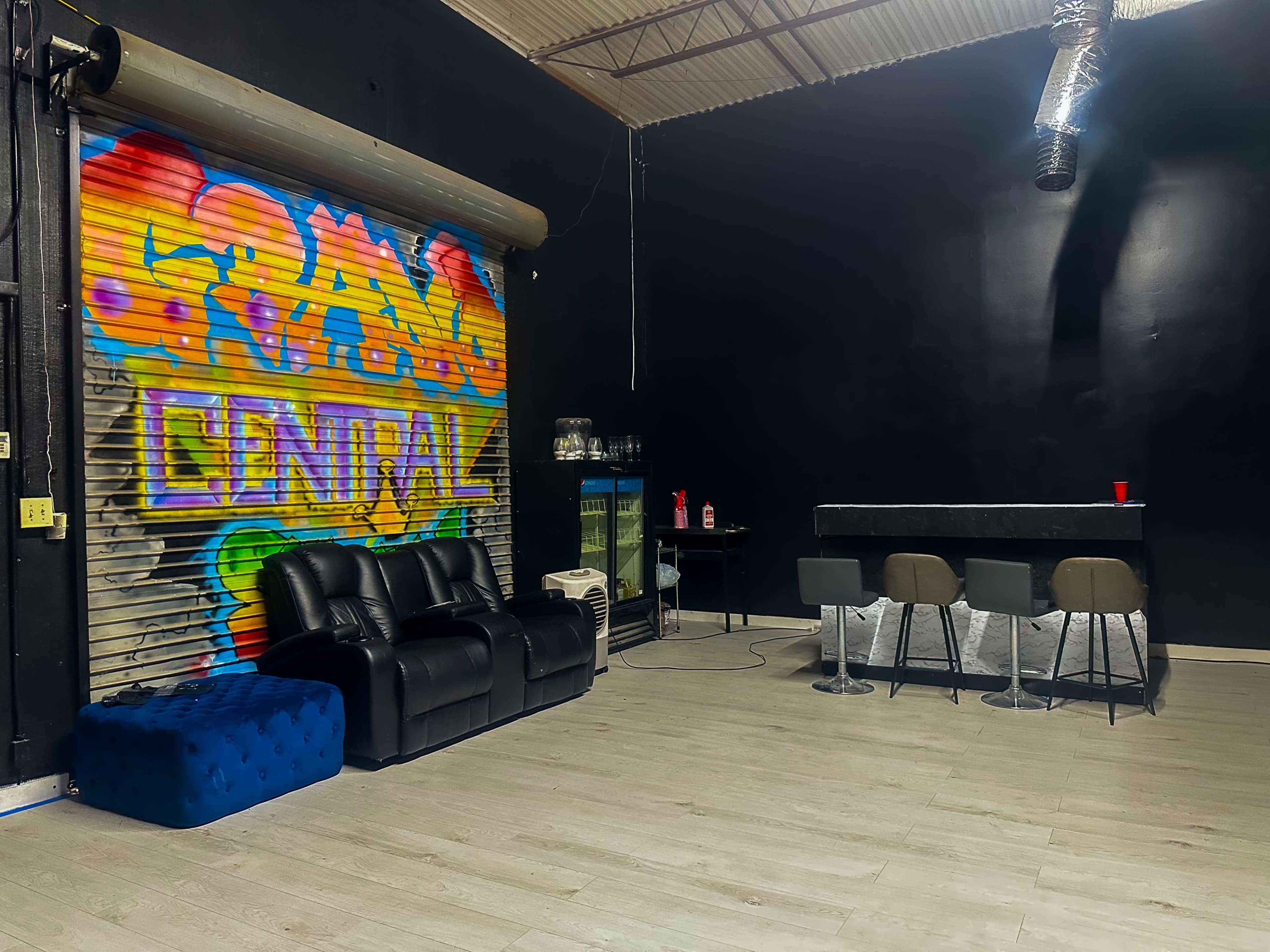 The image shows a spacious room with a colorful graffiti-style mural on one wall, a black bar counter with stools, a blue ottoman, and a black couch.