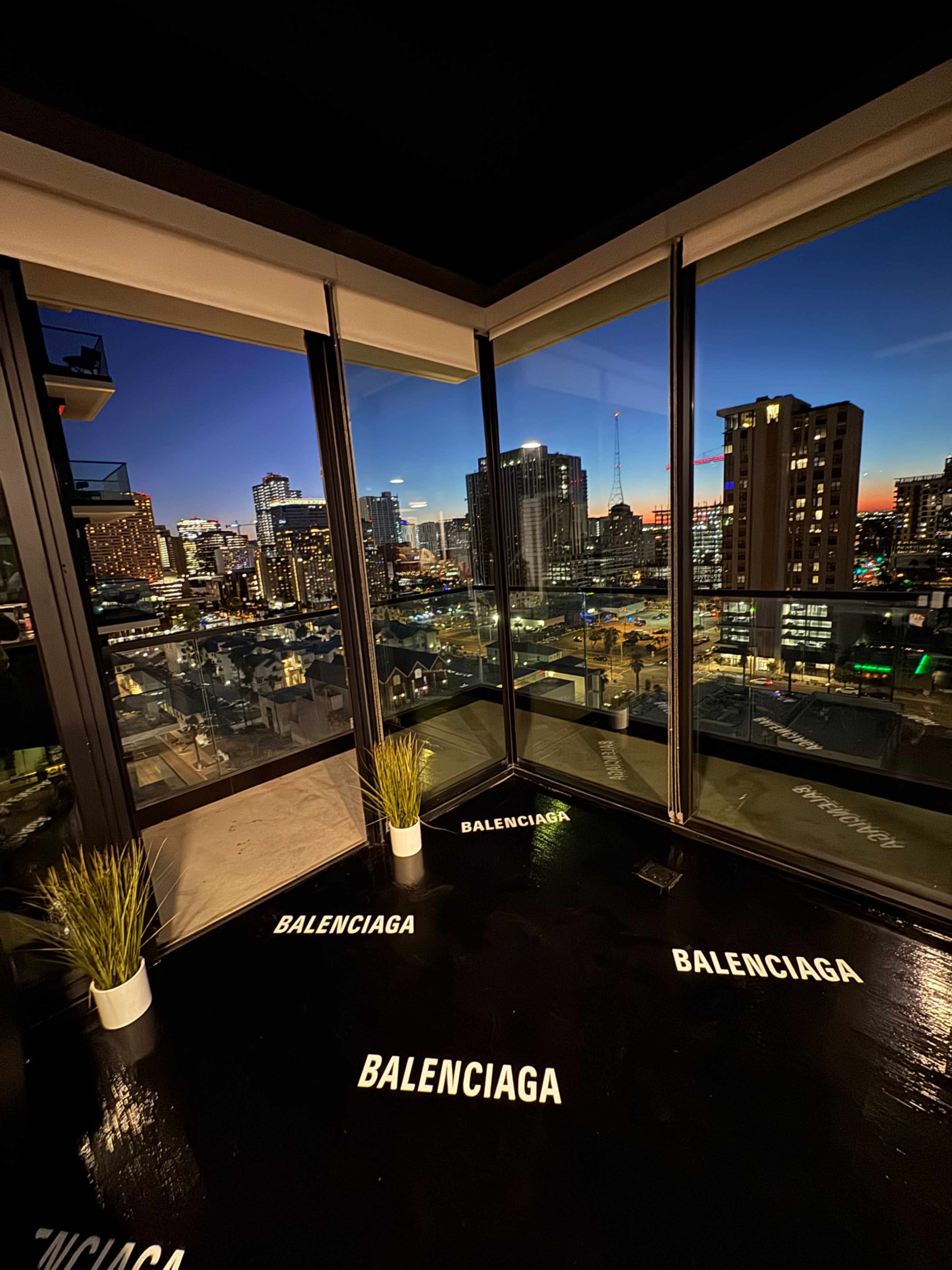 The image shows a modern interior space with large glass windows overlooking a city skyline at dusk, featuring "BALENCIAGA" text displayed on the floor.
