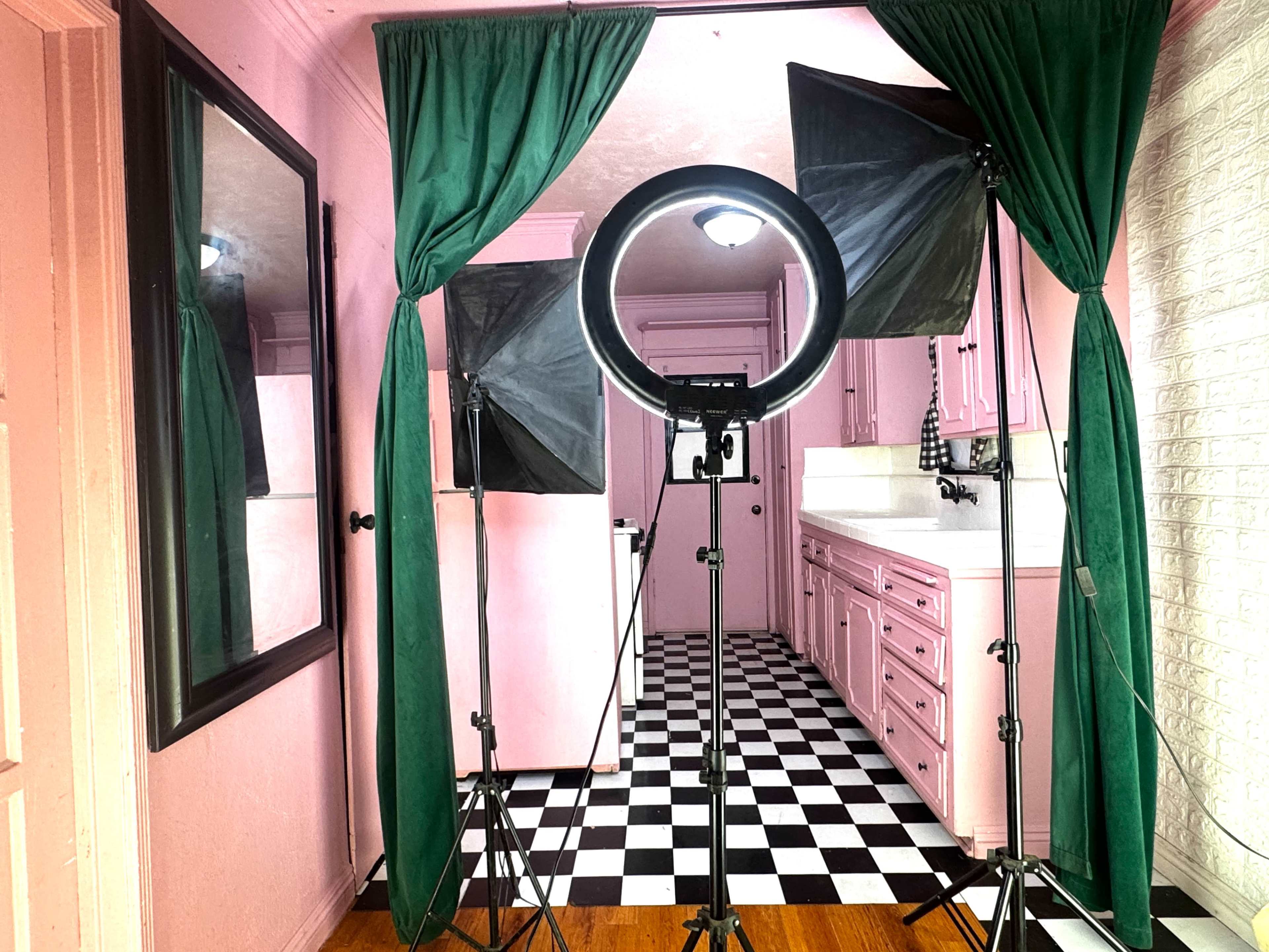 The image shows a room with pink walls, a black and white checkered floor, and two softbox lights set up near a mirror and a kitchen area in the background.