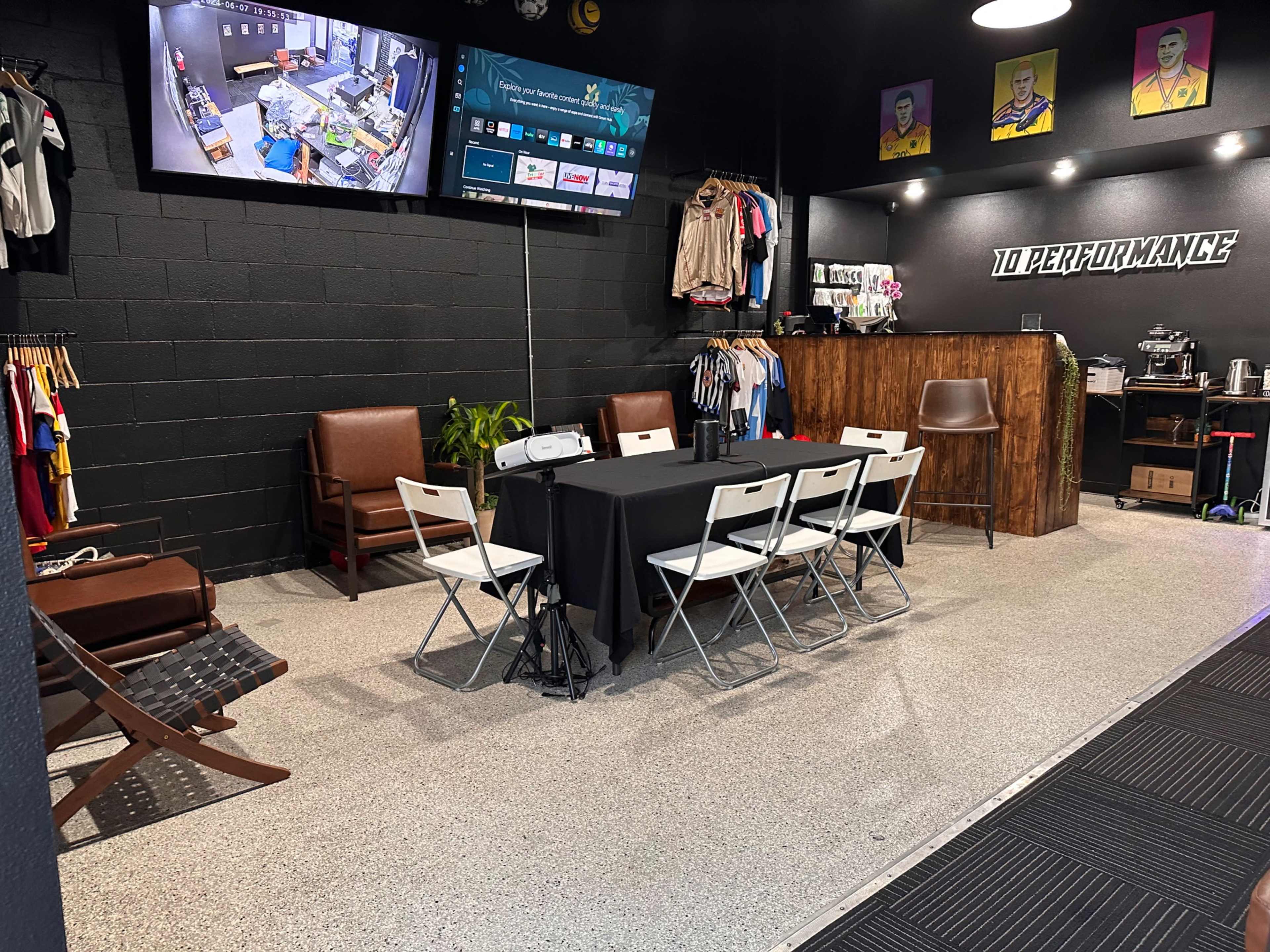 A room with a table and six chairs, a projection screen, clothing racks, and a coffee station, set up for a meeting or presentation.