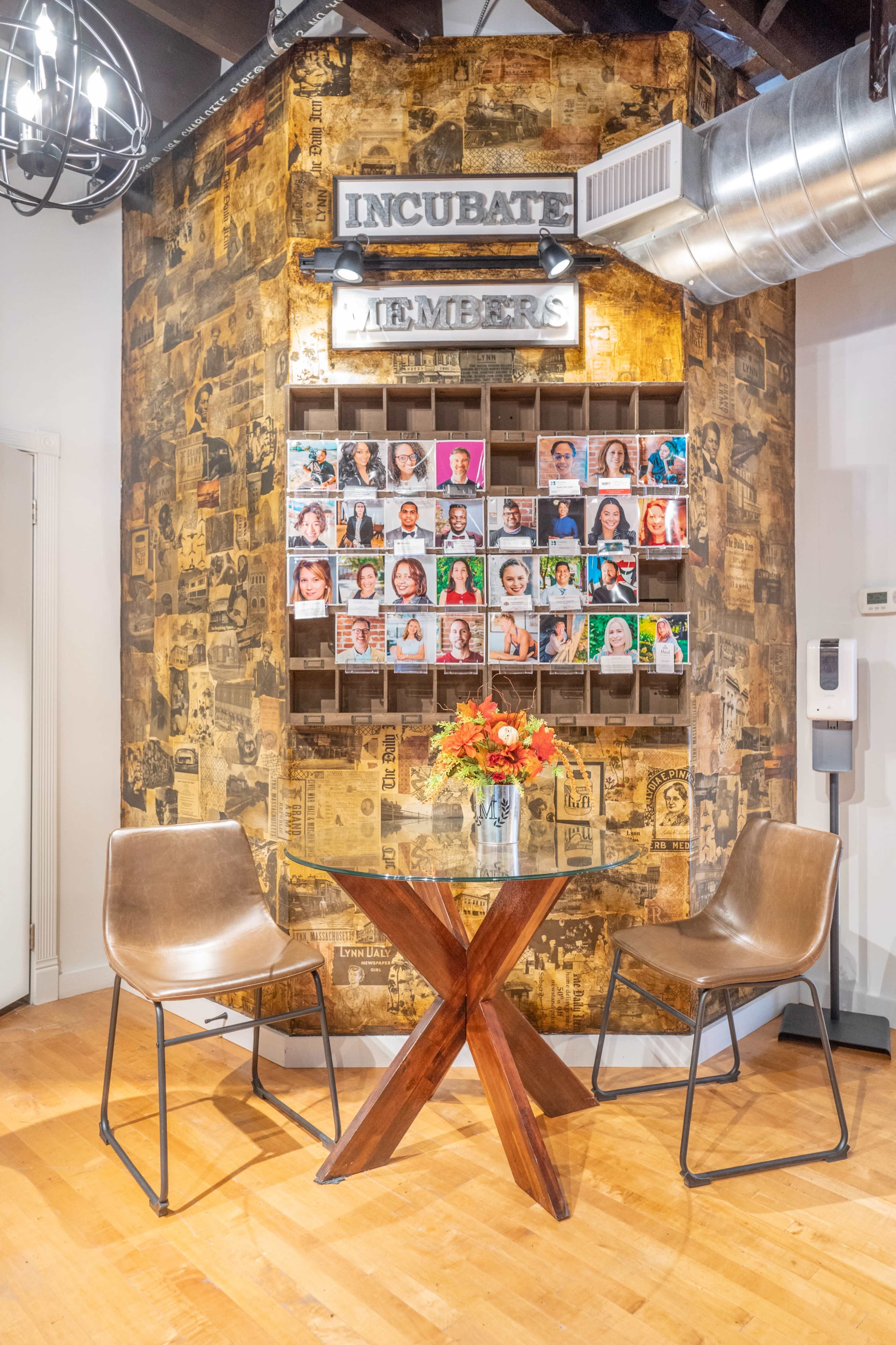 A cozy seating area features a wooden table with two chairs in front of a wall displaying a grid of colorful portraits and the word "INCUBATE" above them.