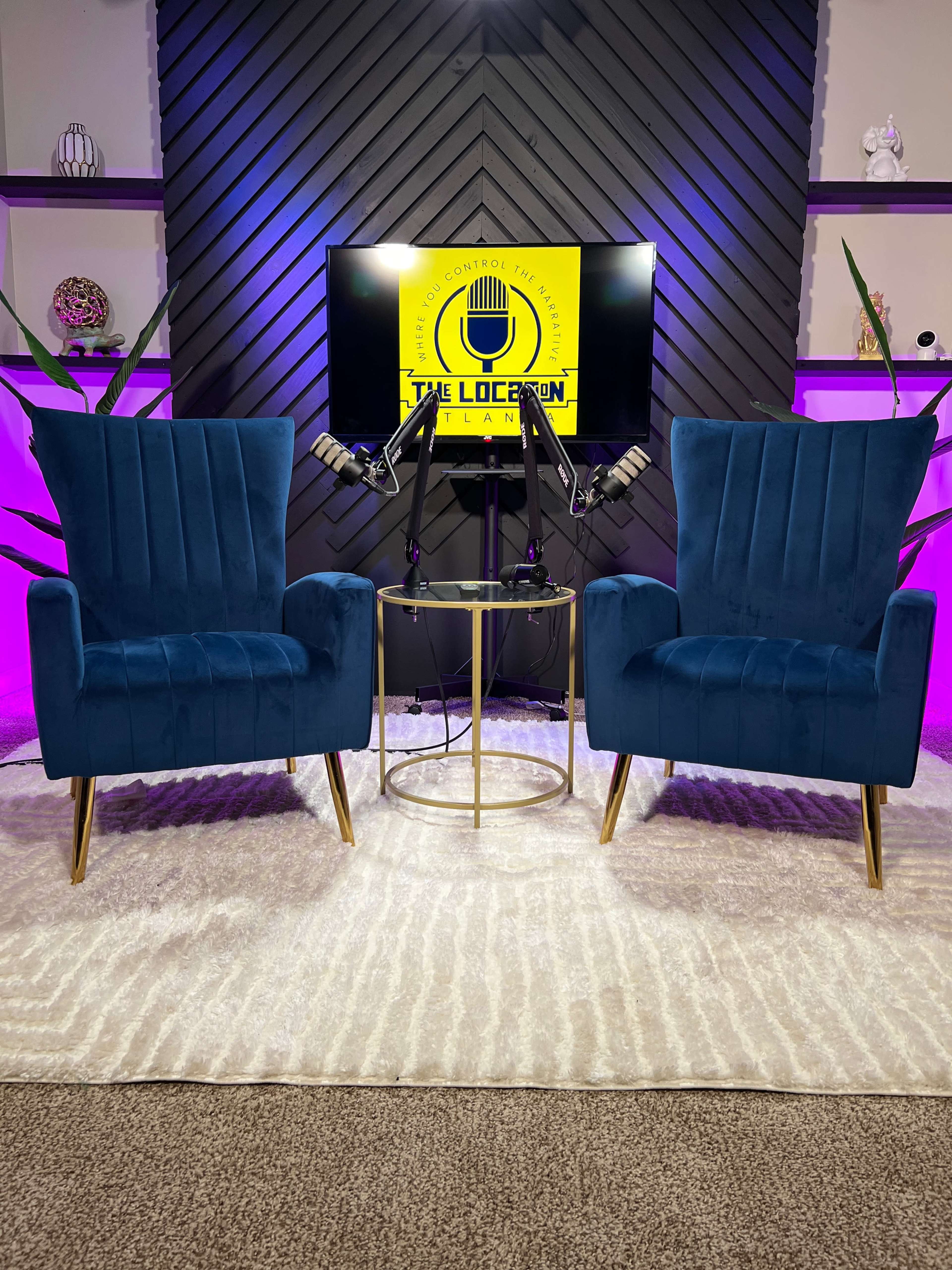 The image shows a small studio setup with two teal chairs facing a circular gold table, a microphone on each chair, and a screen displaying "THE LOUNGE" in the background.