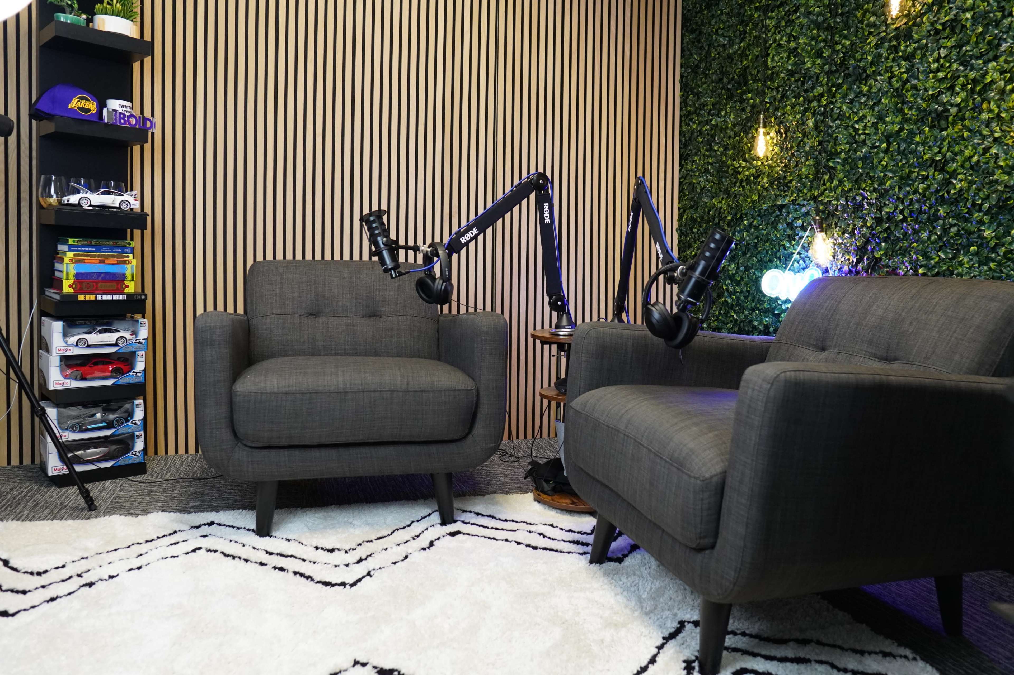 A cozy podcast studio with two gray armchairs, a shelf filled with toys and books, and a backdrop of vertical wooden slats and green plants.