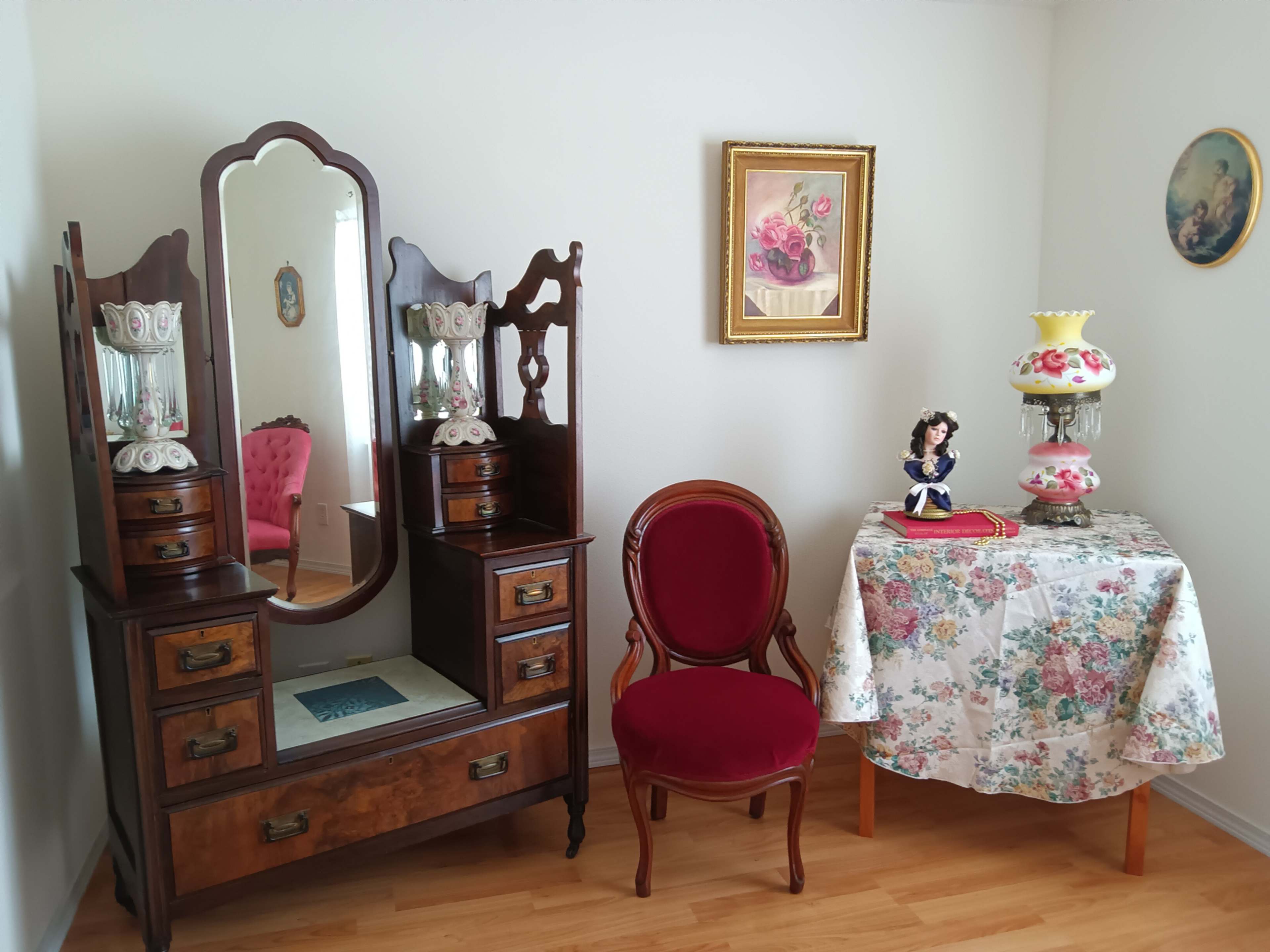 The image shows a vintage room setting with a wooden dresser featuring a large mirror, a red upholstered chair, and a small table adorned with a floral tablecloth, a decorative porcelain doll, and a lamp.
