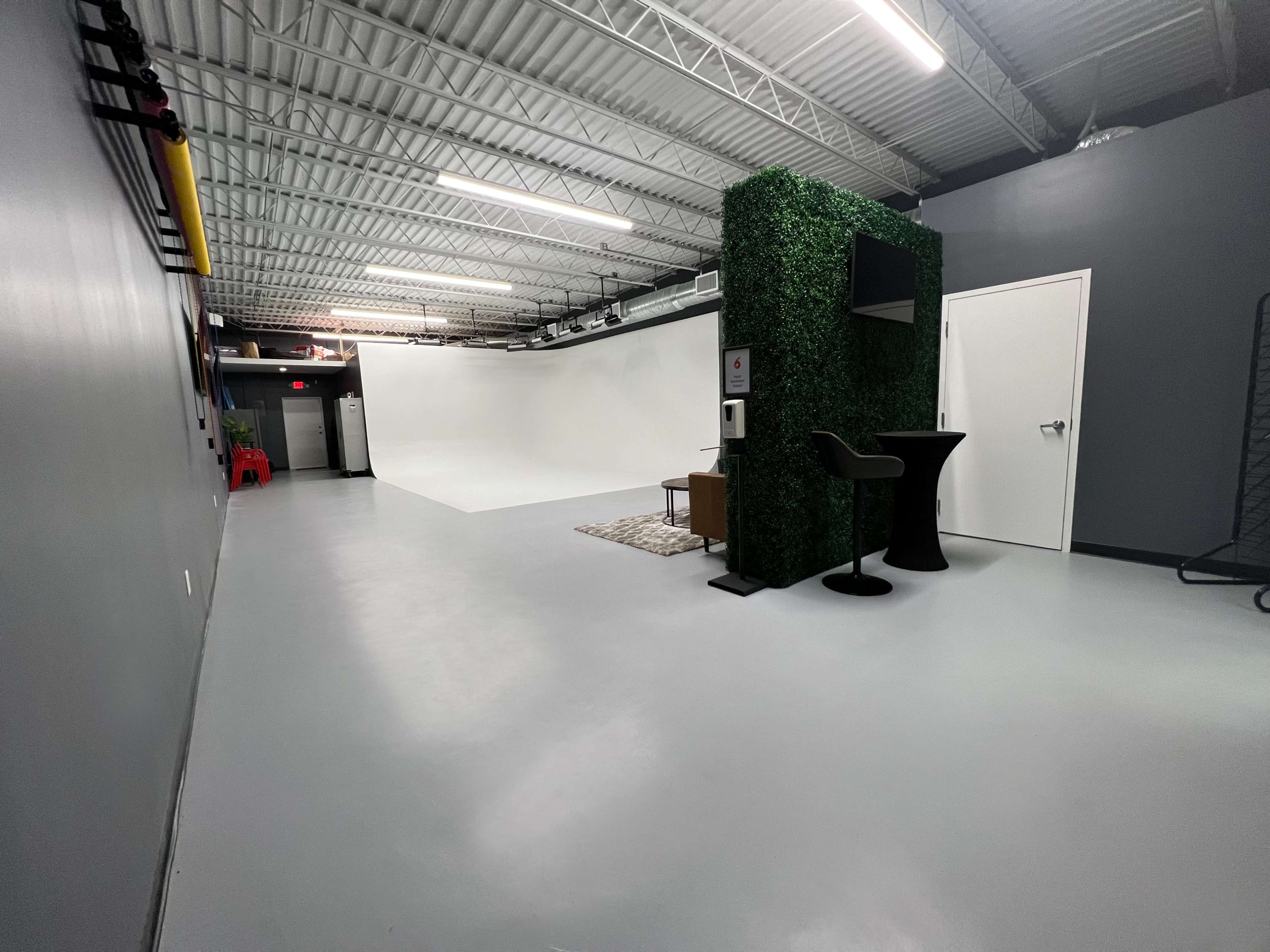 The image shows an empty photography studio featuring a white backdrop, a small seating area, a table, and a green wall section.