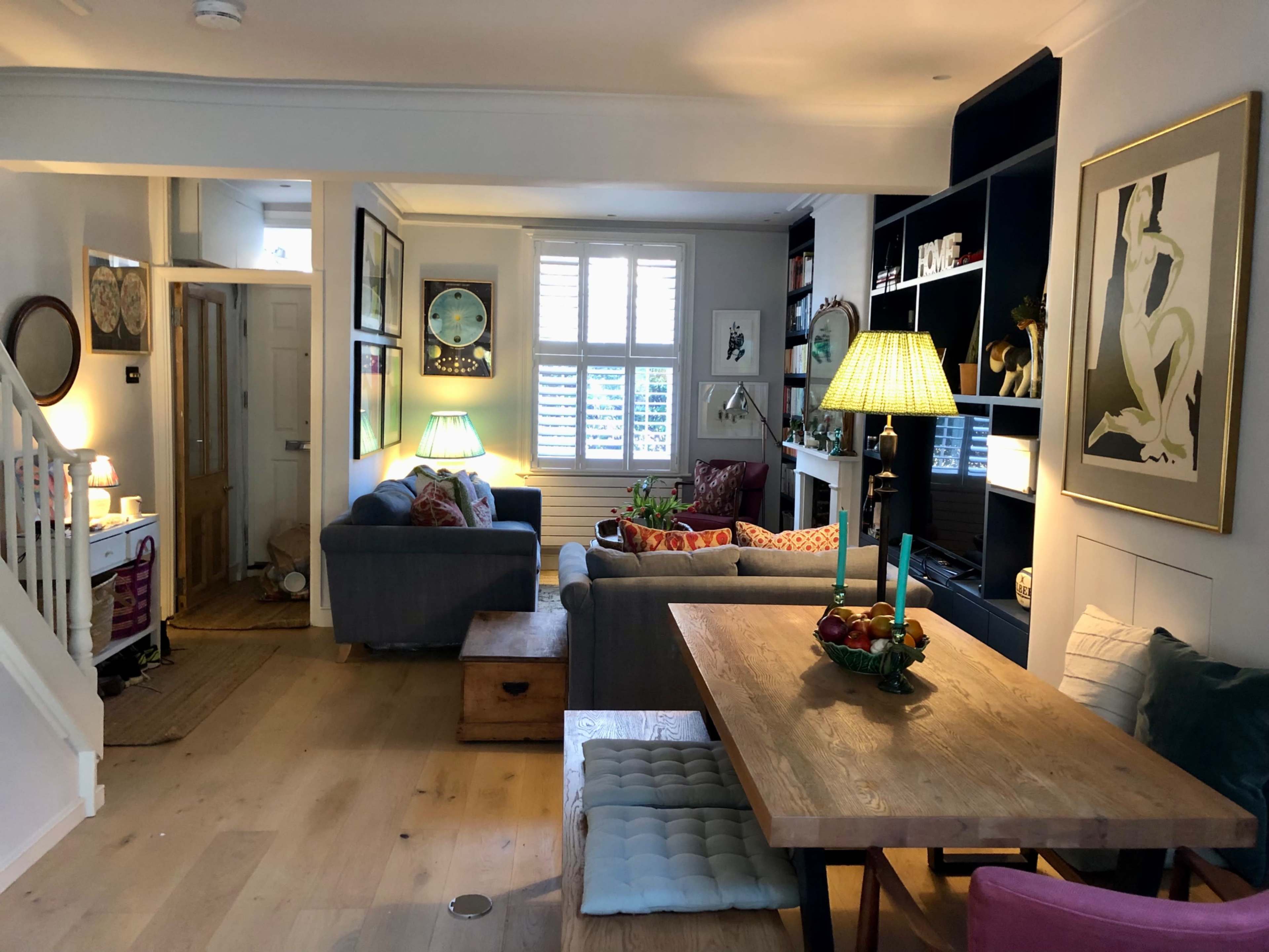 The image shows a cozy living and dining area with a wooden table, comfortable seating, art on the walls, and a staircase leading to another room.