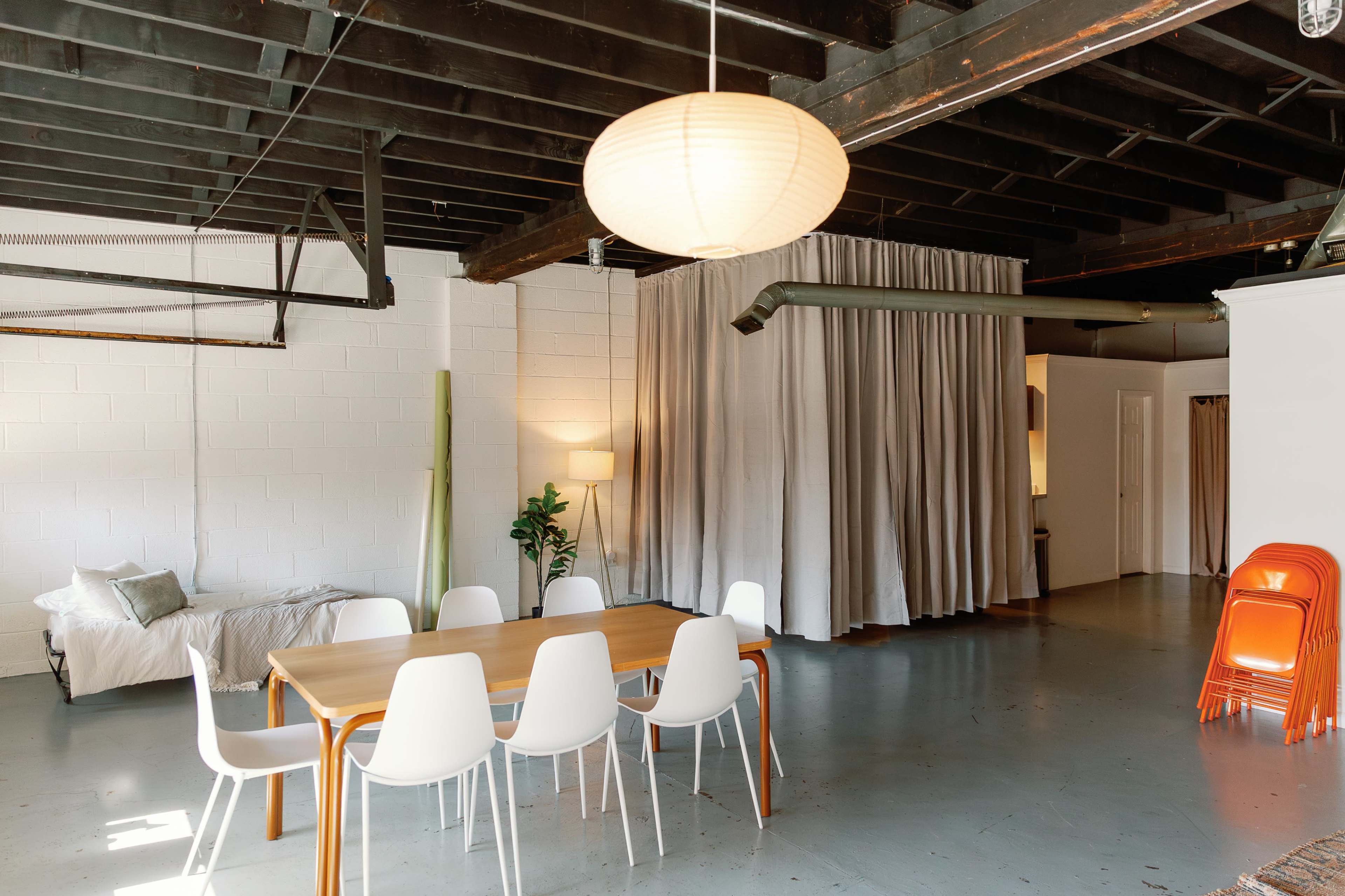 The image shows a modern open space featuring a wooden dining table surrounded by white chairs, with a bed in the background and curtains dividing different areas of the room.