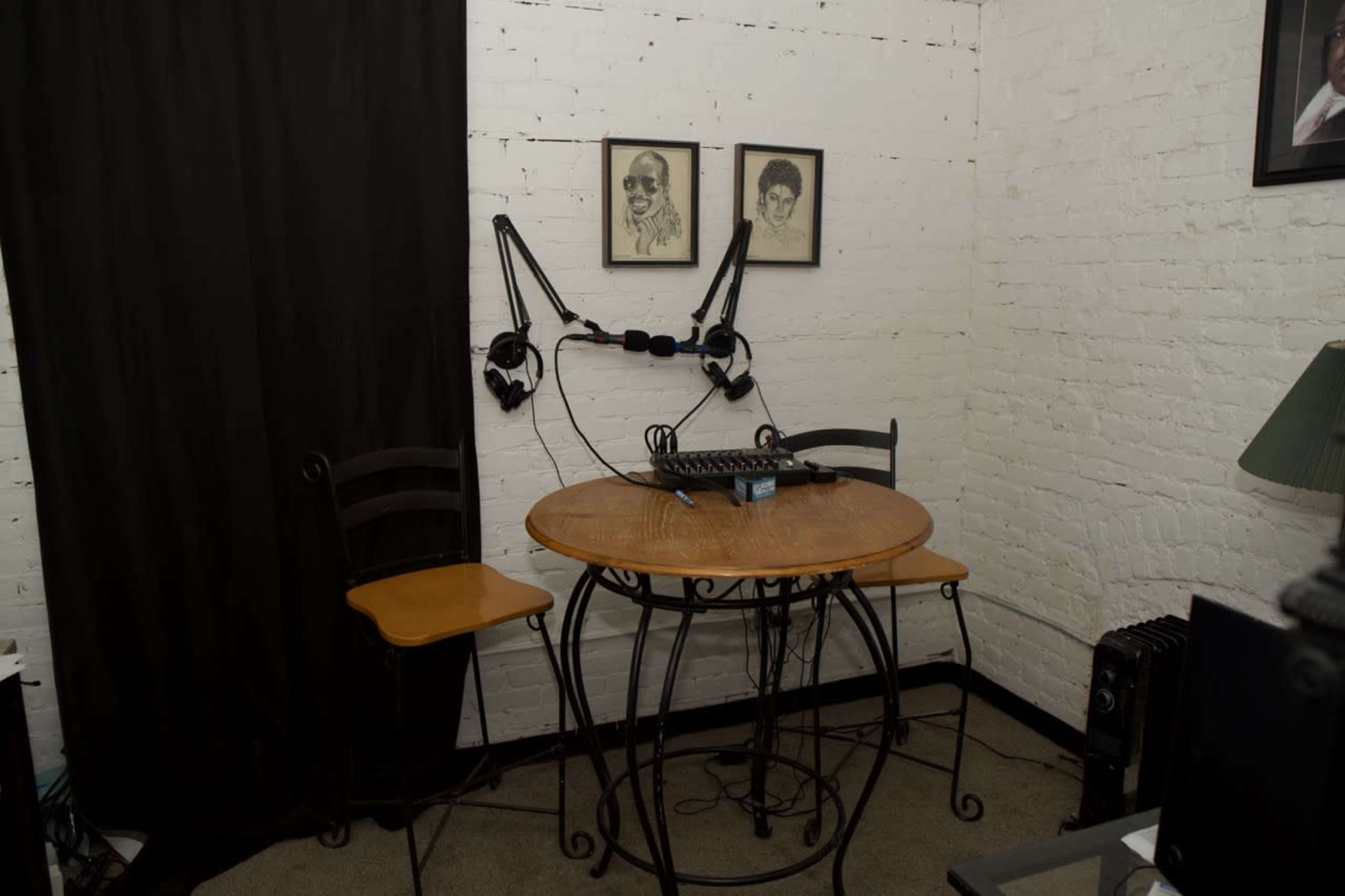 The image shows a small recording studio setup with a round table, two chairs, and microphones mounted on adjustable arms attached to the wall.