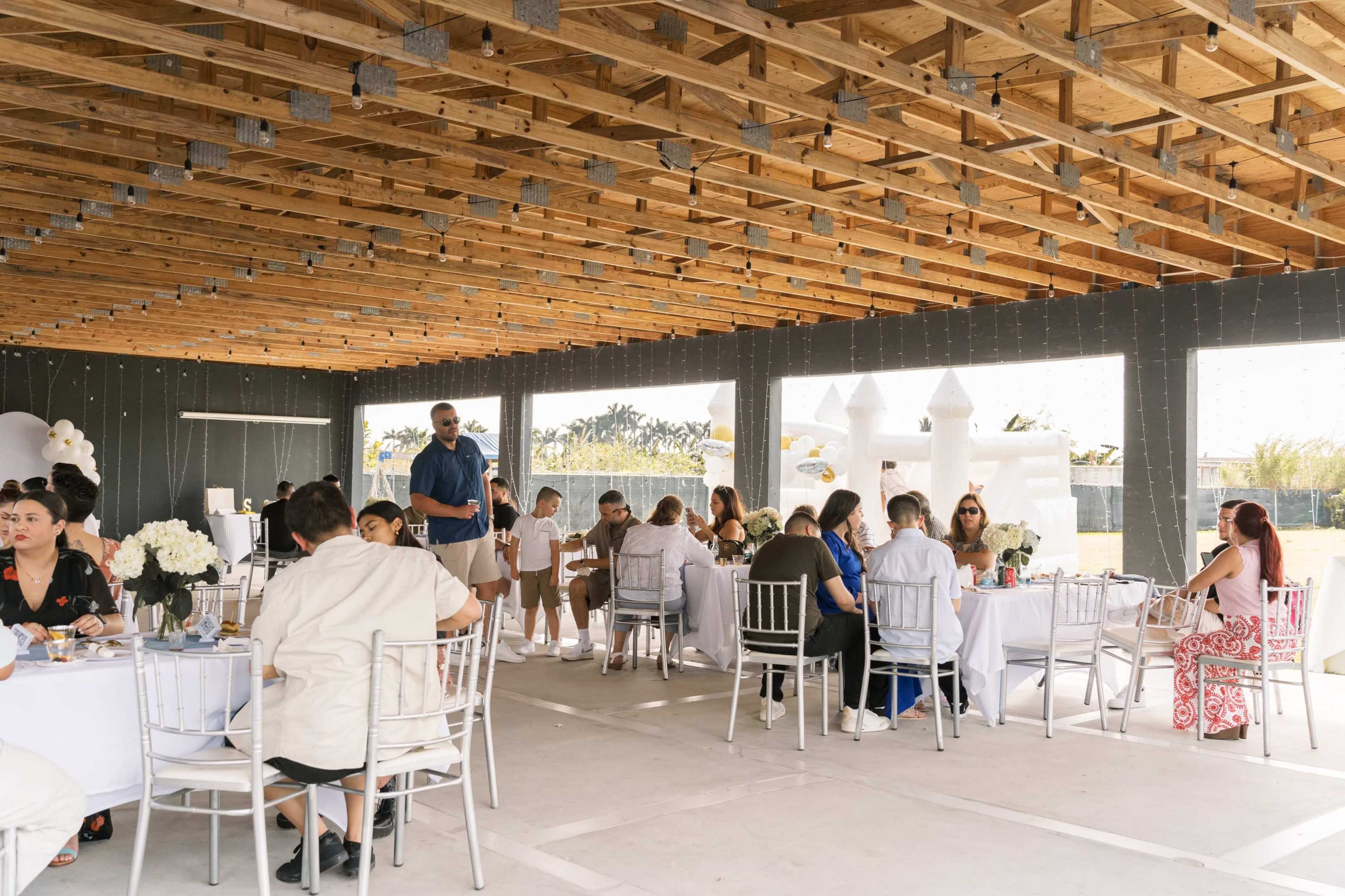 A group of people sits at round tables in a spacious, partially enclosed venue decorated for an event.