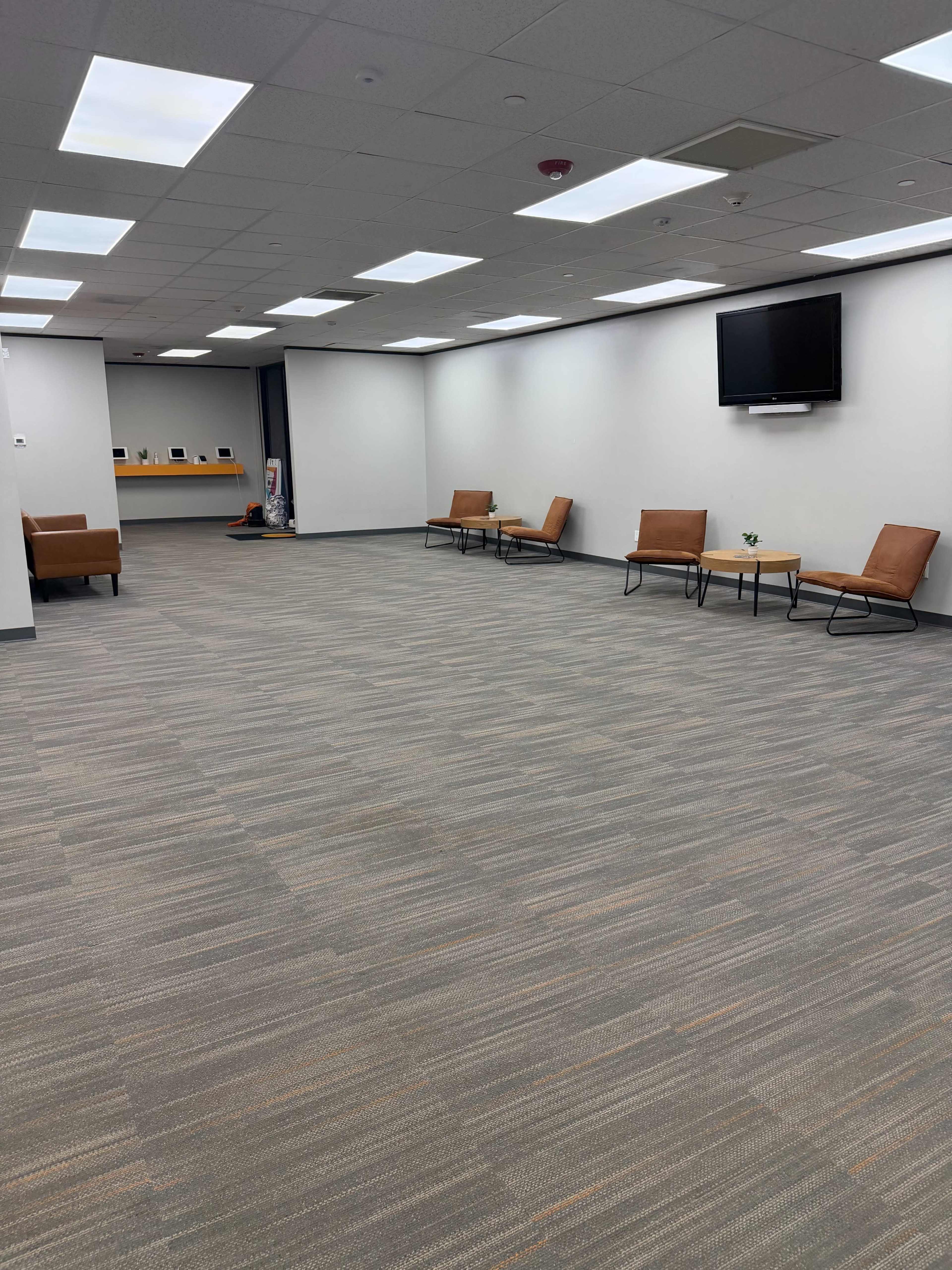 The image shows an empty office waiting area with brown chairs, small tables, and a mounted television on the wall.