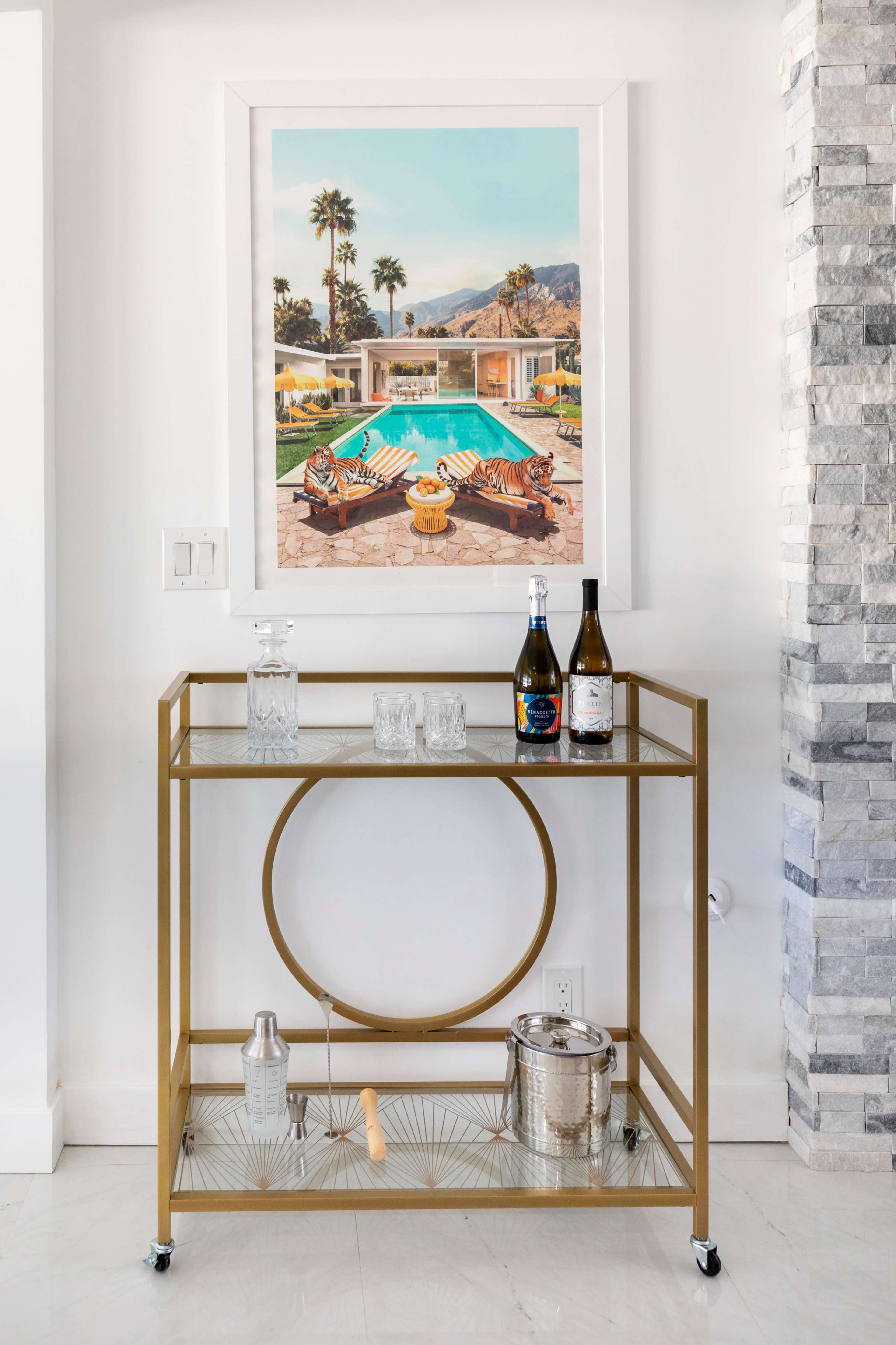 A stylish bar cart with glassware, bottles of wine, and an art piece of a poolside scene is placed against a white wall with stone accents.