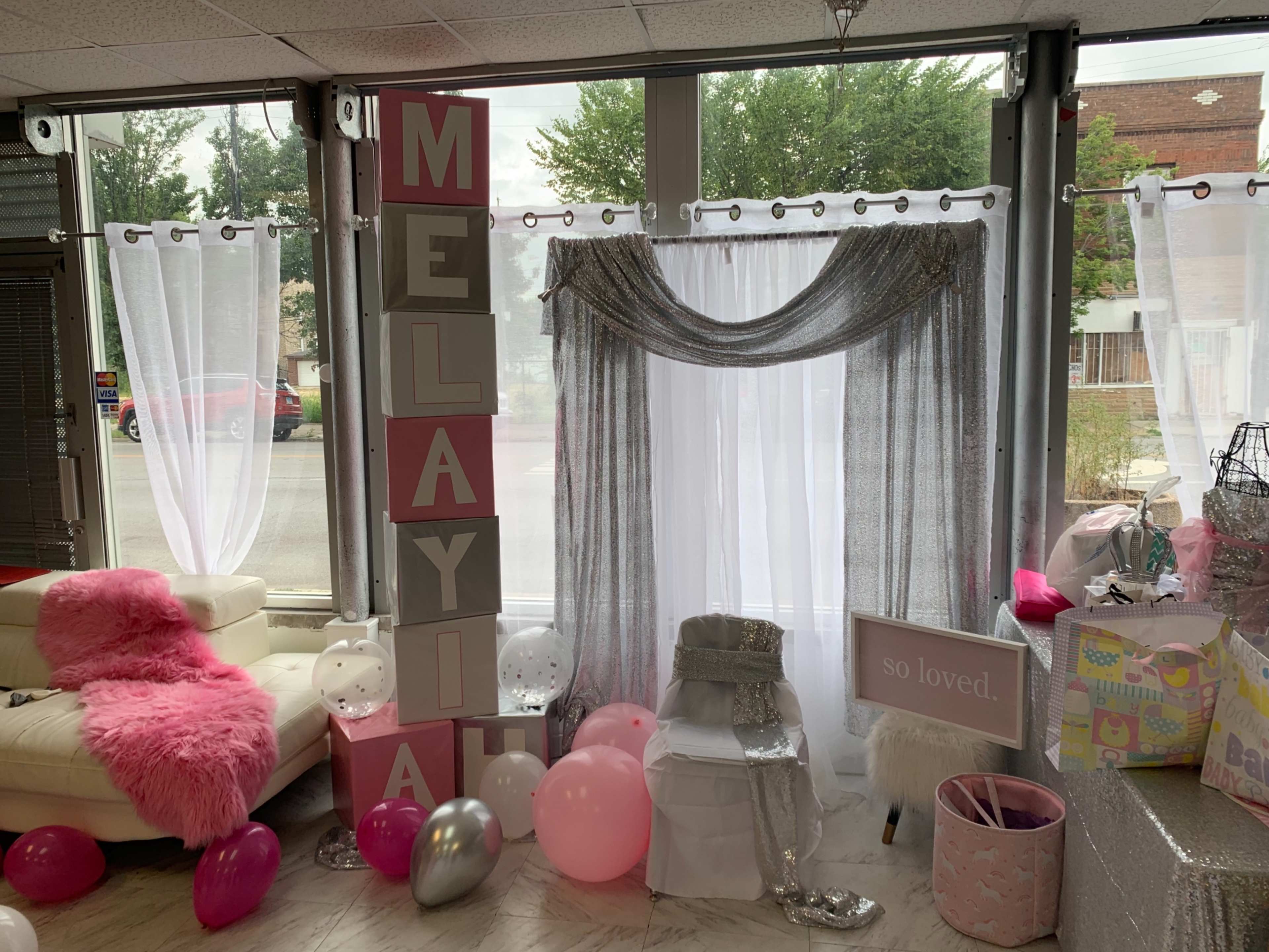 A decorated space for a celebration, featuring large letters spelling "MELAYIAH," sheer white drapes, a silver-accented backdrop, and various party decorations including balloons and gifts.